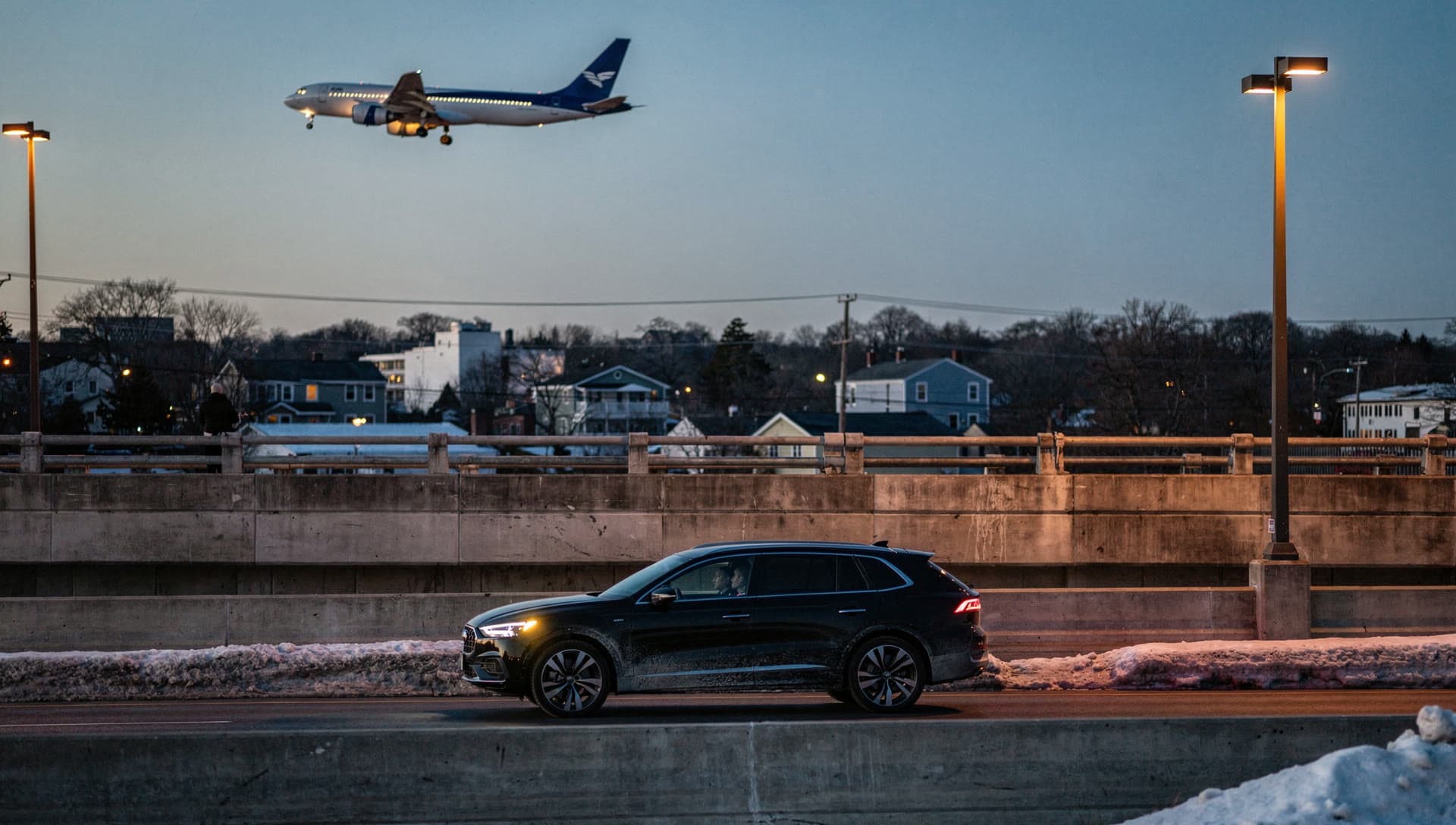 A black executive SUV crossing an empty airport access overpass at blue hour, an aircraft on final approach with its landing lights on above the roadway.