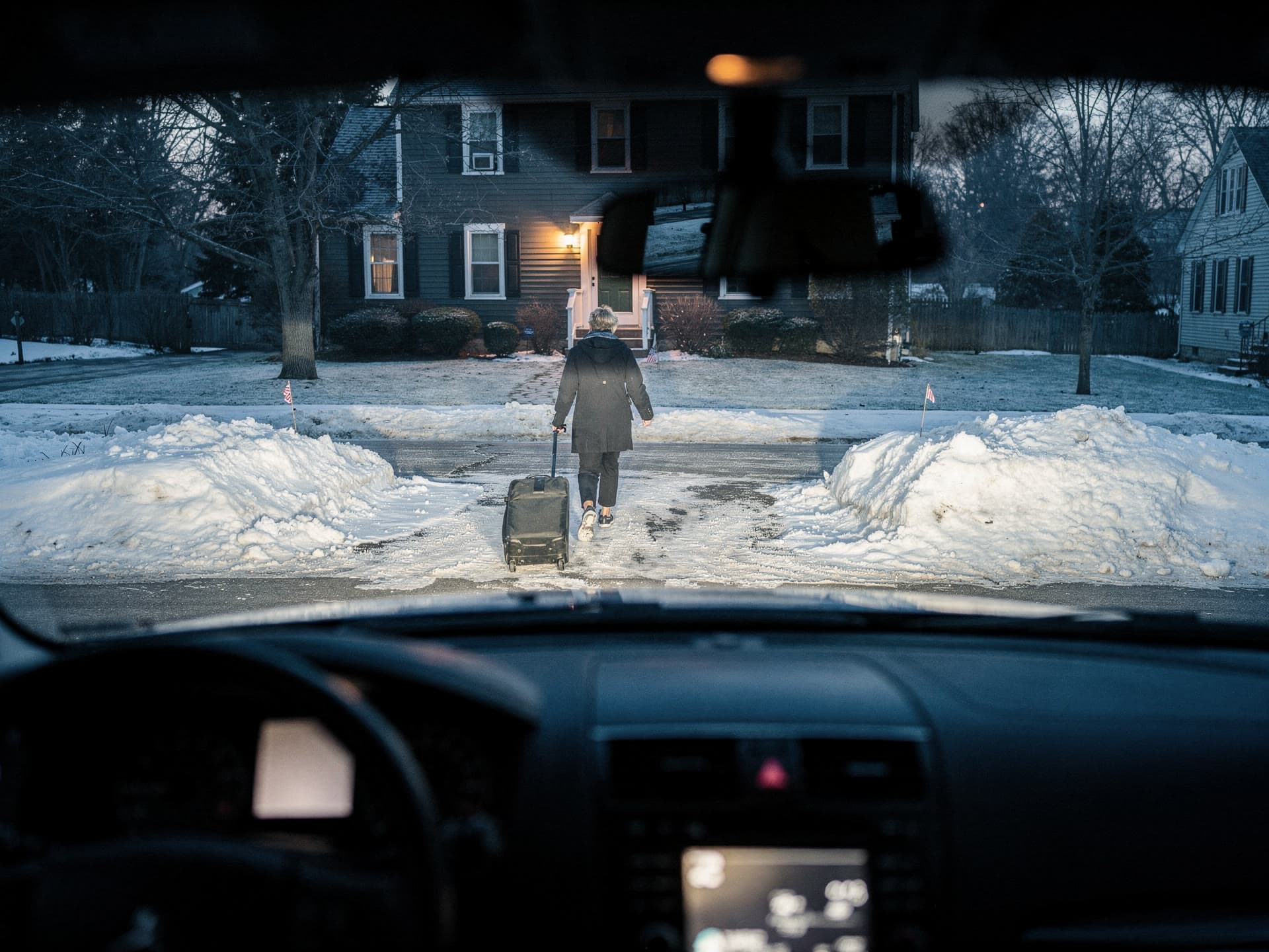 The view through a windshield from the driver's seat as a traveller walks down her own front path with a roller bag, porch light behind her.