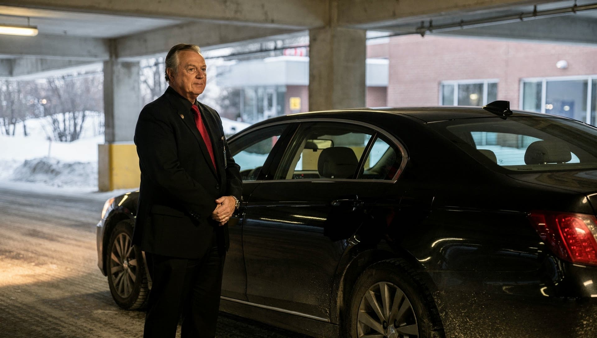 A chauffeur in a black suit and red tie stands with his hands clasped beside a black sedan in a concrete parking garage, with snow outside.