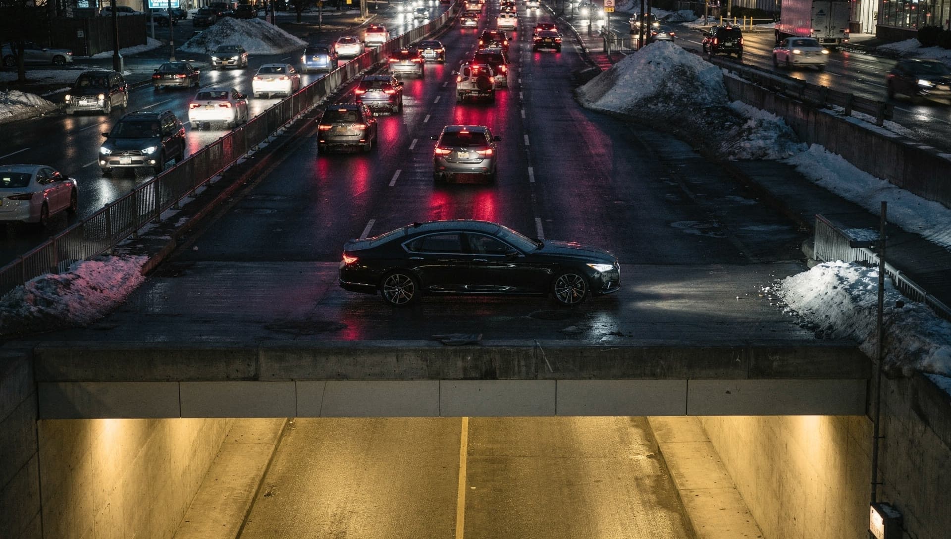 An elevated dusk view of a wet multi lane highway with red taillights and plowed snowbanks, a black sedan alone on the deck above a lit tunnel portal.