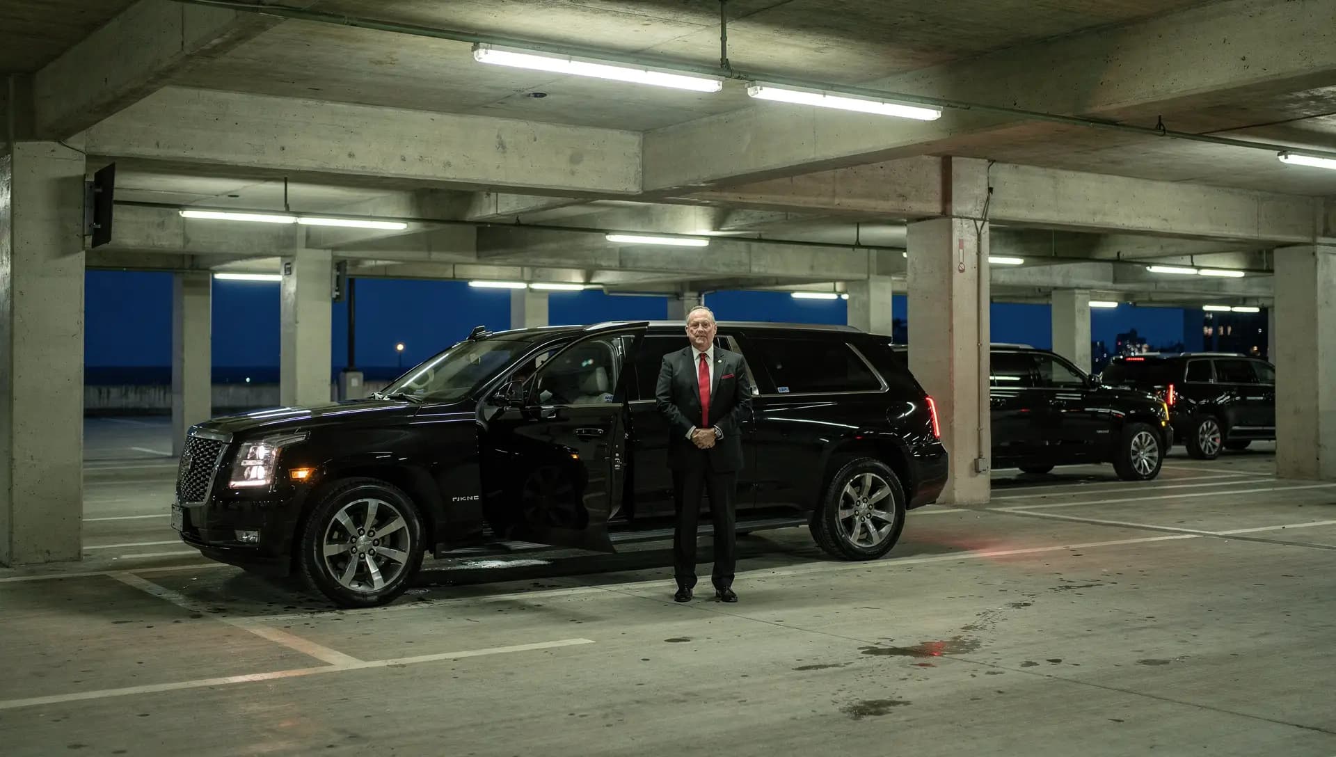 A chauffeur waiting beside a black SUV at the limousine stand inside the Logan Airport parking garage before dawn