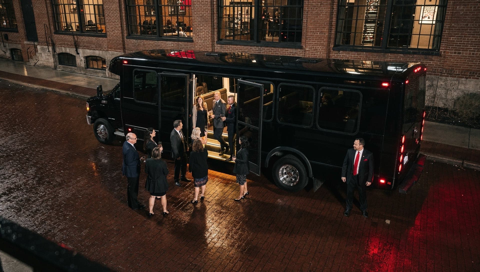 An elevated view of passengers gathering and boarding a black party bus on a wet brick street outside a brick building.