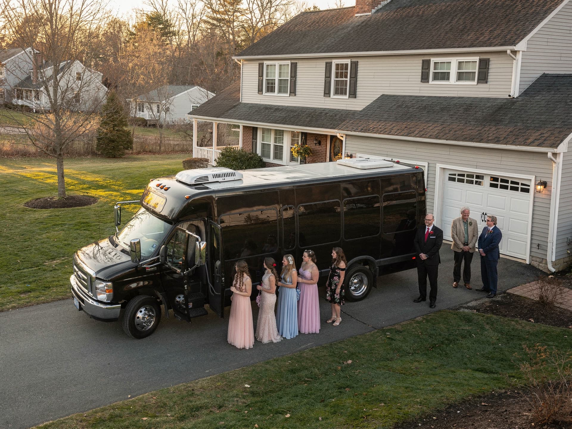 A group of teenagers in formalwear boarding a party bus in a driveway while two parents stand watching from beside the garage.