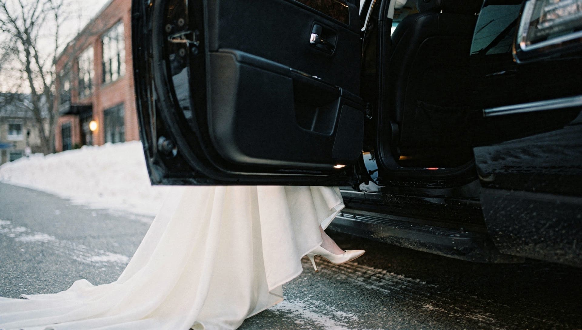 A low ground level view of an ivory gown hem and a satin heel stepping from the open rear door of a black vehicle onto wet pavement.
