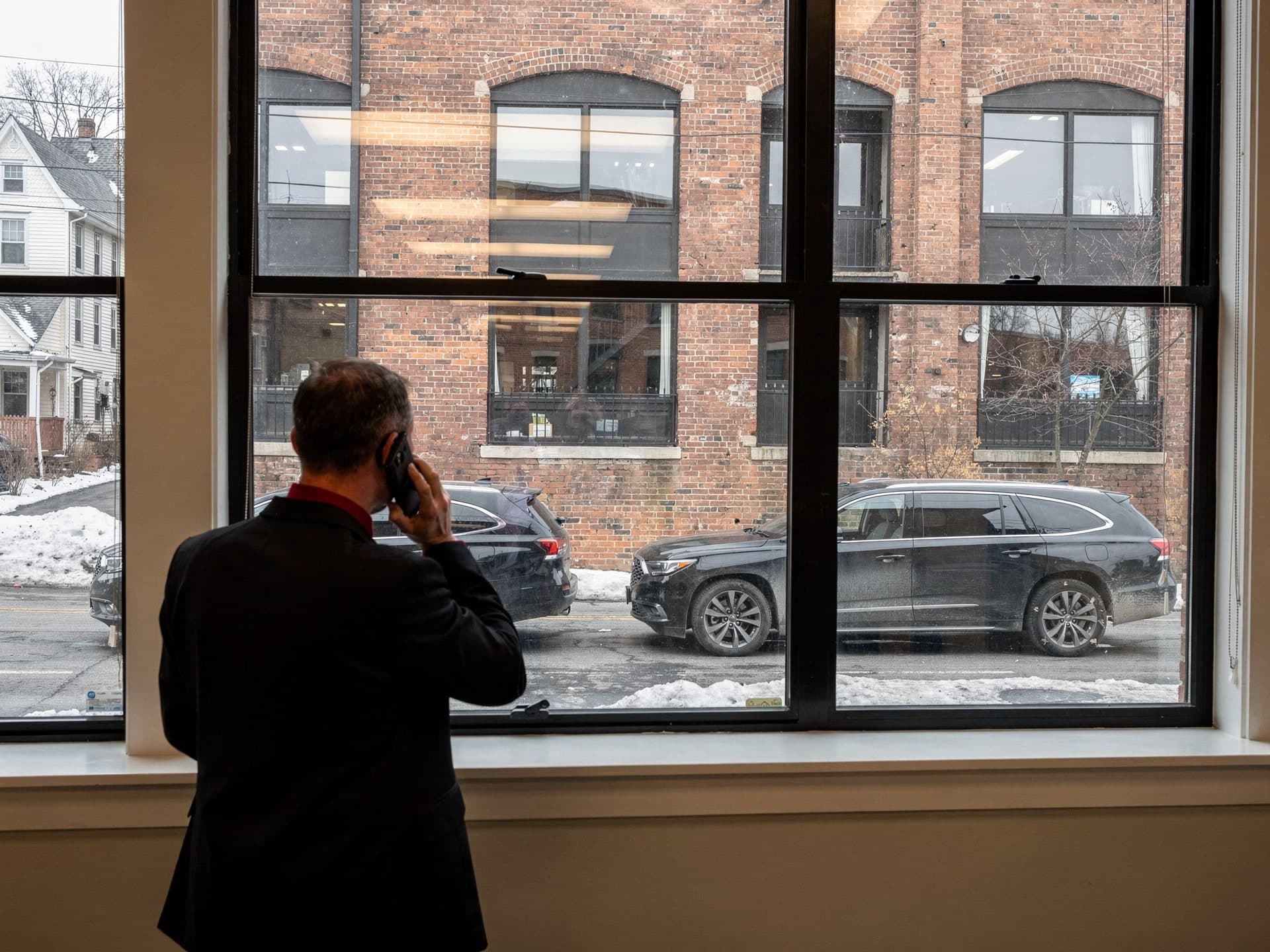 Seen from behind, a man in a dark suit and red tie holds a phone to his ear at a tall office window, looking out at two black SUVs parked along a snow-lined street beneath a brick mill building.