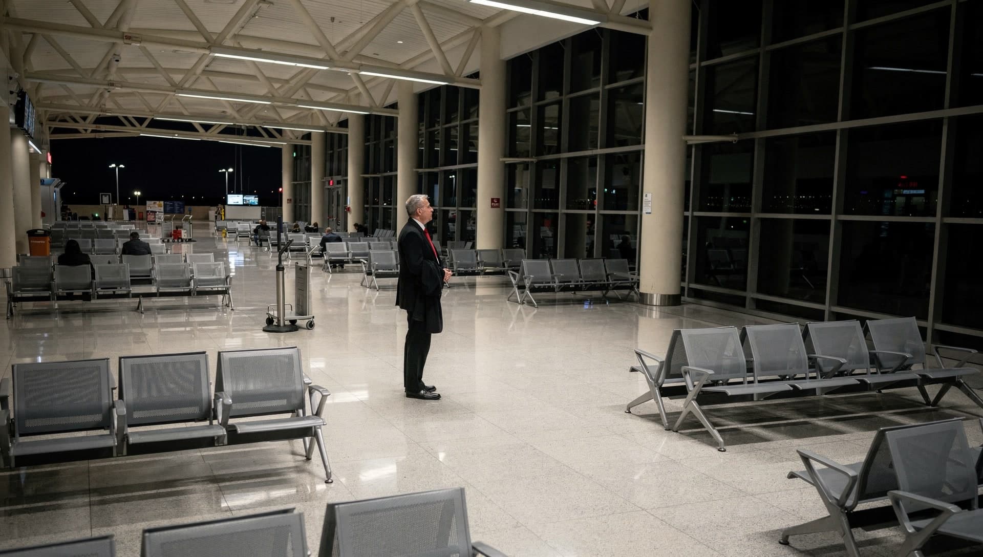 A chauffeur in a black suit and red tie stands alone in a near-empty airport terminal hall late at night, hands clasped in front of him, surrounded by rows of empty grey seating with dark glass beyond.