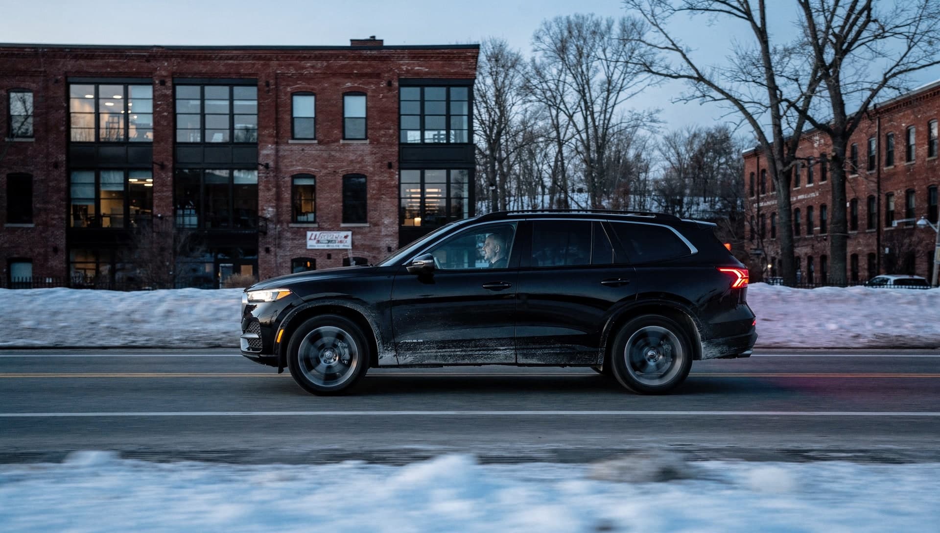 A black SUV drives past a restored brick mill building on a two-lane road at dusk, its wheels blurred with motion and plowed snow banked along both sides of the road.