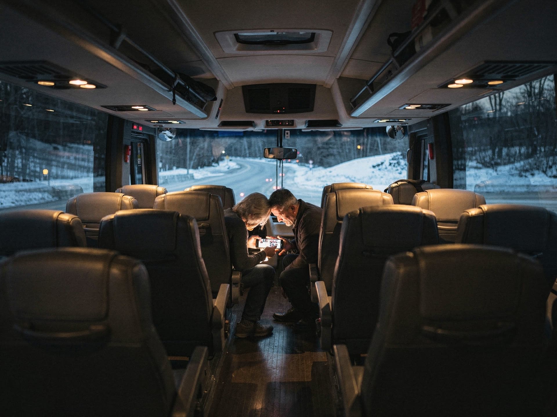 The center aisle of a motorcoach in the evening, two riders in high back seats leaning together over a phone.