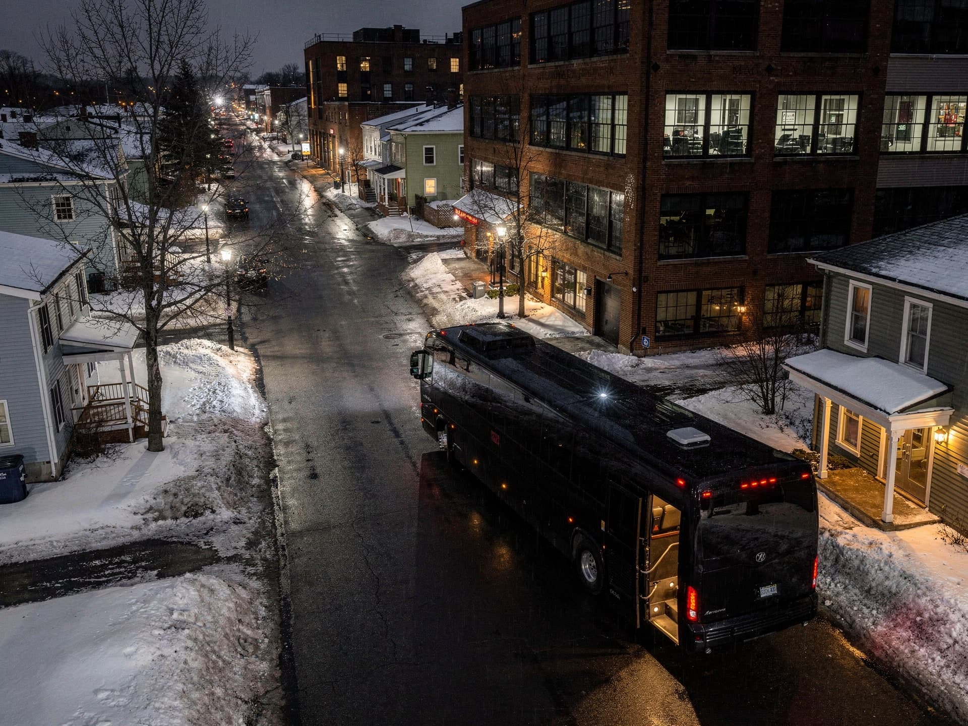 Seen from above on a wet winter night, a black motorcoach stands at the kerb of a lit mill street with its passenger door open and the step glowing, nobody in frame.