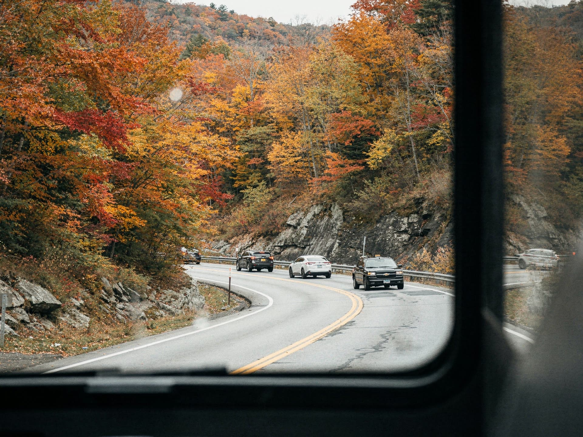 A mountain highway curve seen from a high coach window, red and gold maples filling the frame on both sides.