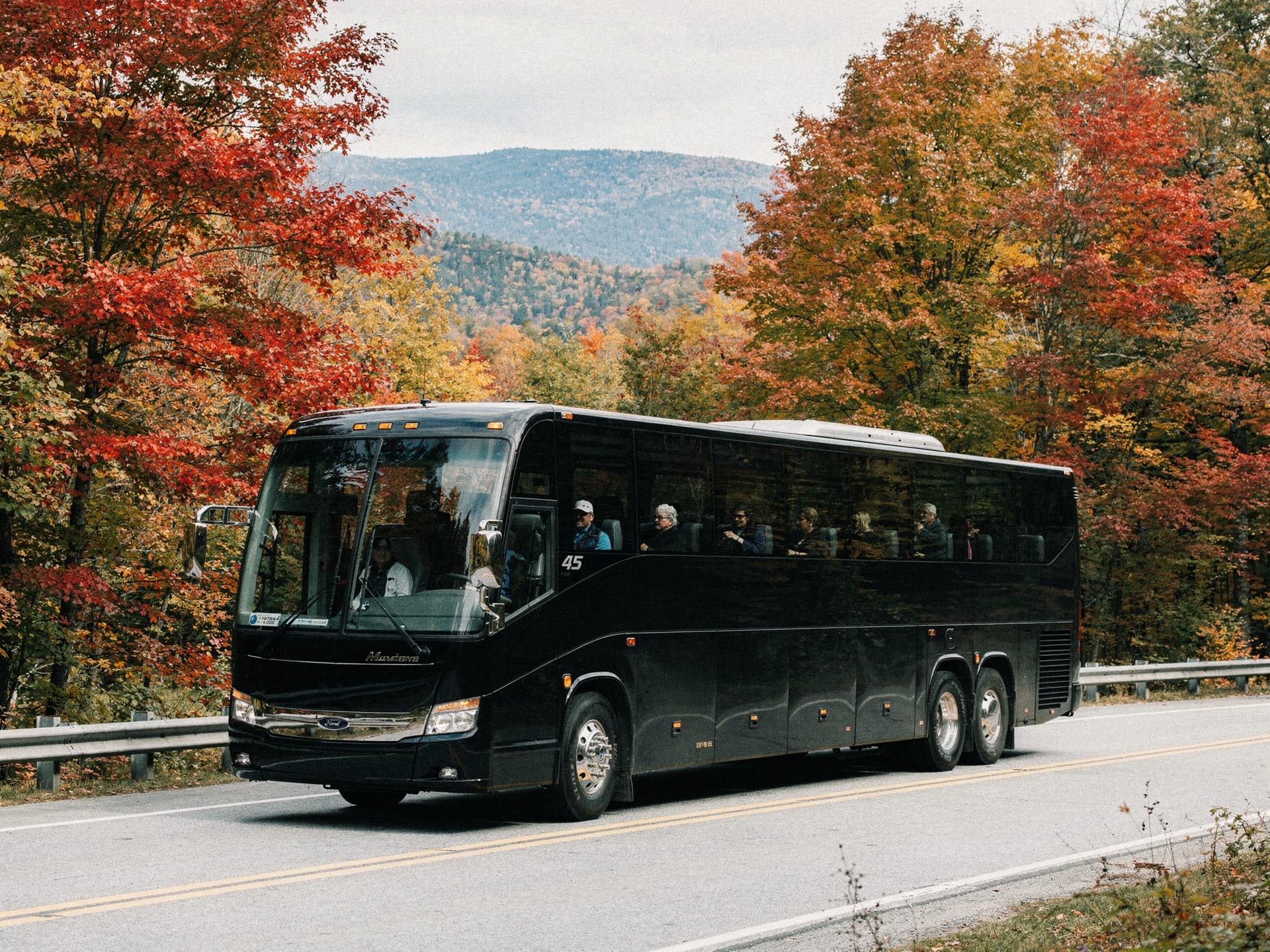 A motorcoach on a mountain highway framed by red and gold maples, passengers visible at the windows looking out at the ridgeline.