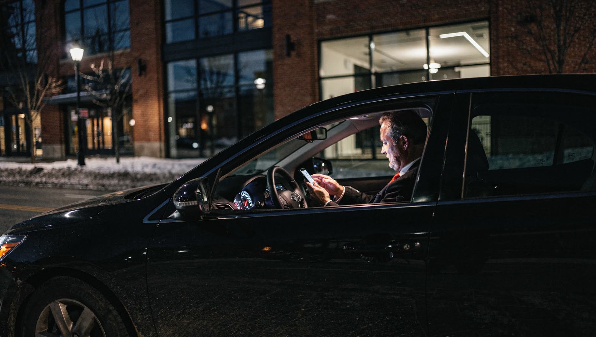 A chauffeur sitting in a parked car at night with the interior light on, seen from outside through the driver's window