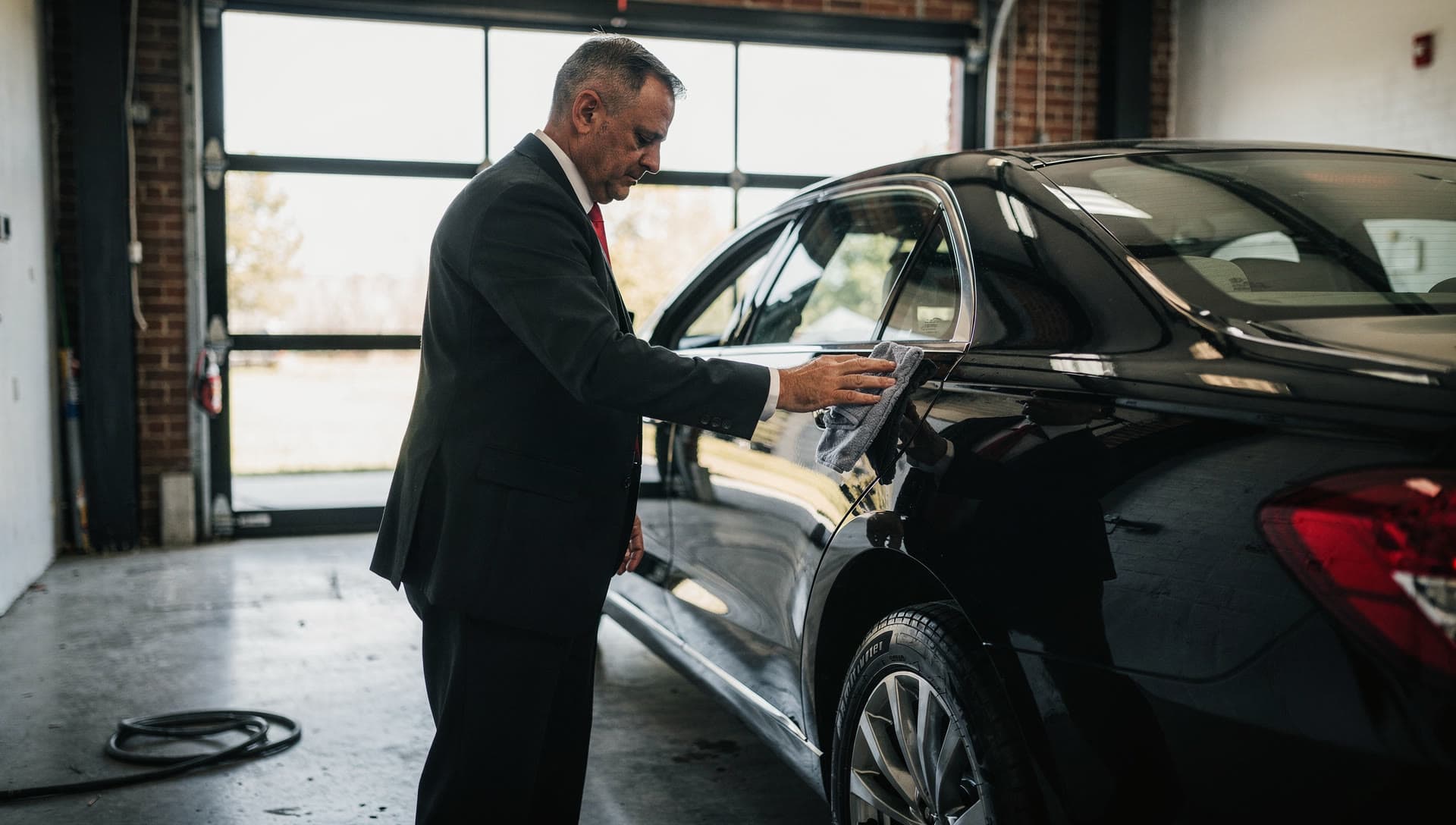 A chauffeur detailing the rear quarter panel of a black vehicle in a garage bay with the roll up door open behind him, the reflection sharp in the paint.