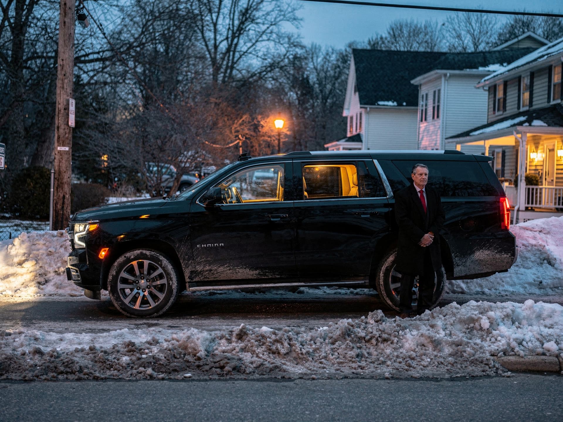 A black Chevrolet Tahoe on a snow edged suburban street at dusk, warm interior light showing through the windows.