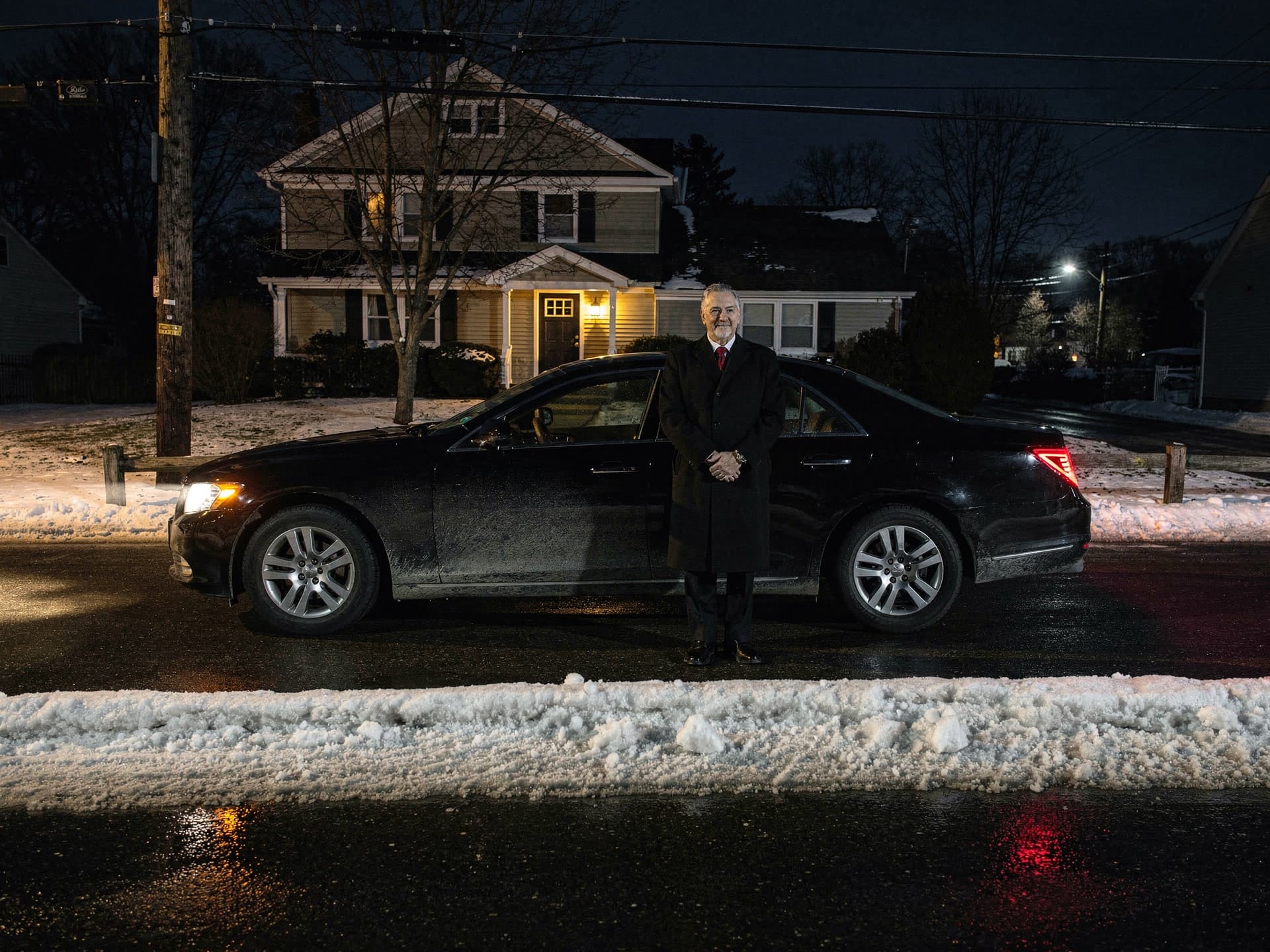 A silver haired chauffeur in a long black overcoat and red tie standing at the rear door of an unmarked black executive sedan parked on a snowed in residential street before dawn, its headlights on and a porch lamp lit at the clapboard house behind him.
