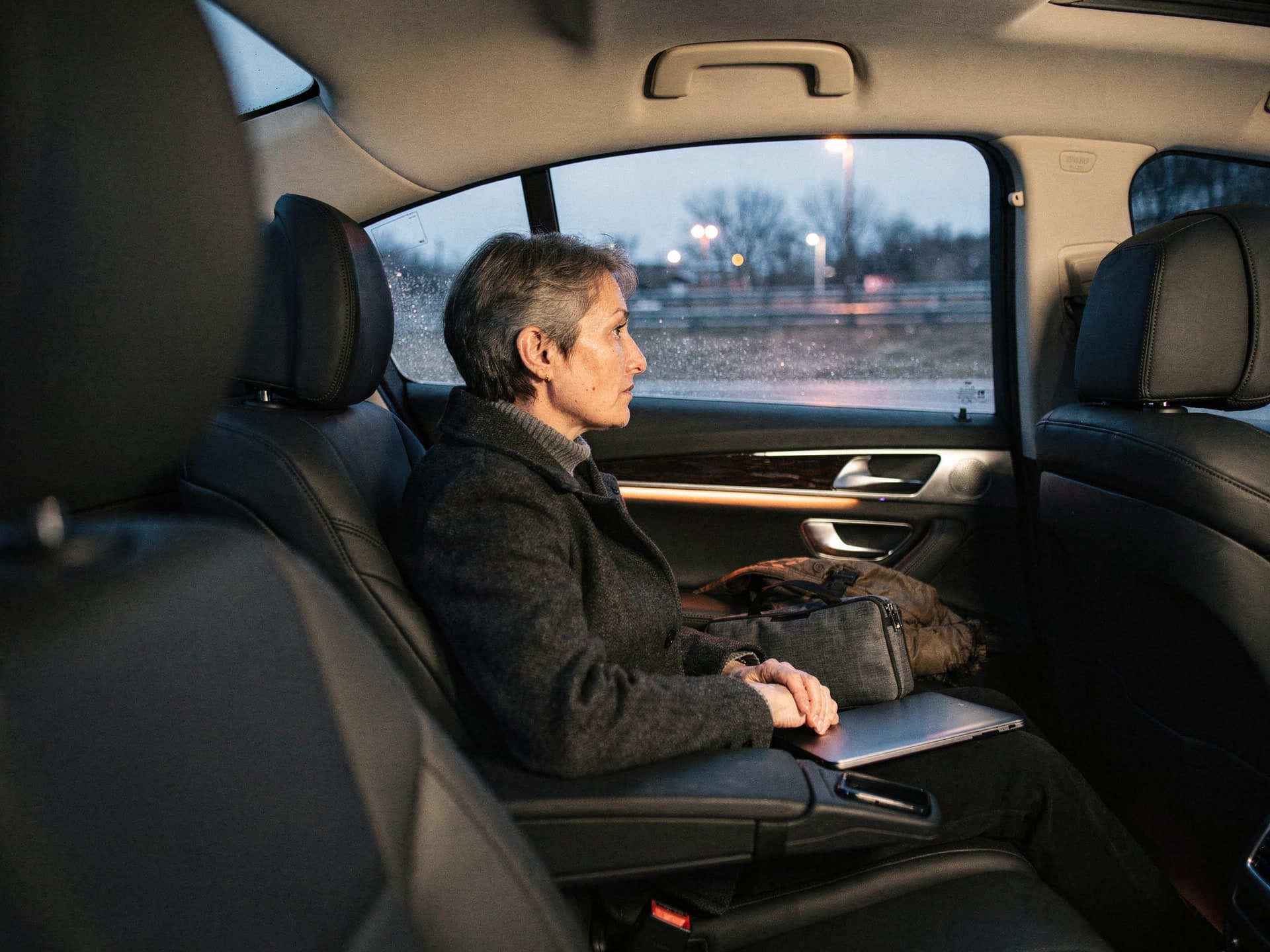 A woman in her fifties riding in the rear seat of a black executive sedan at dusk, a closed laptop and a slim case resting on her lap, looking out of the window at the highway going past.