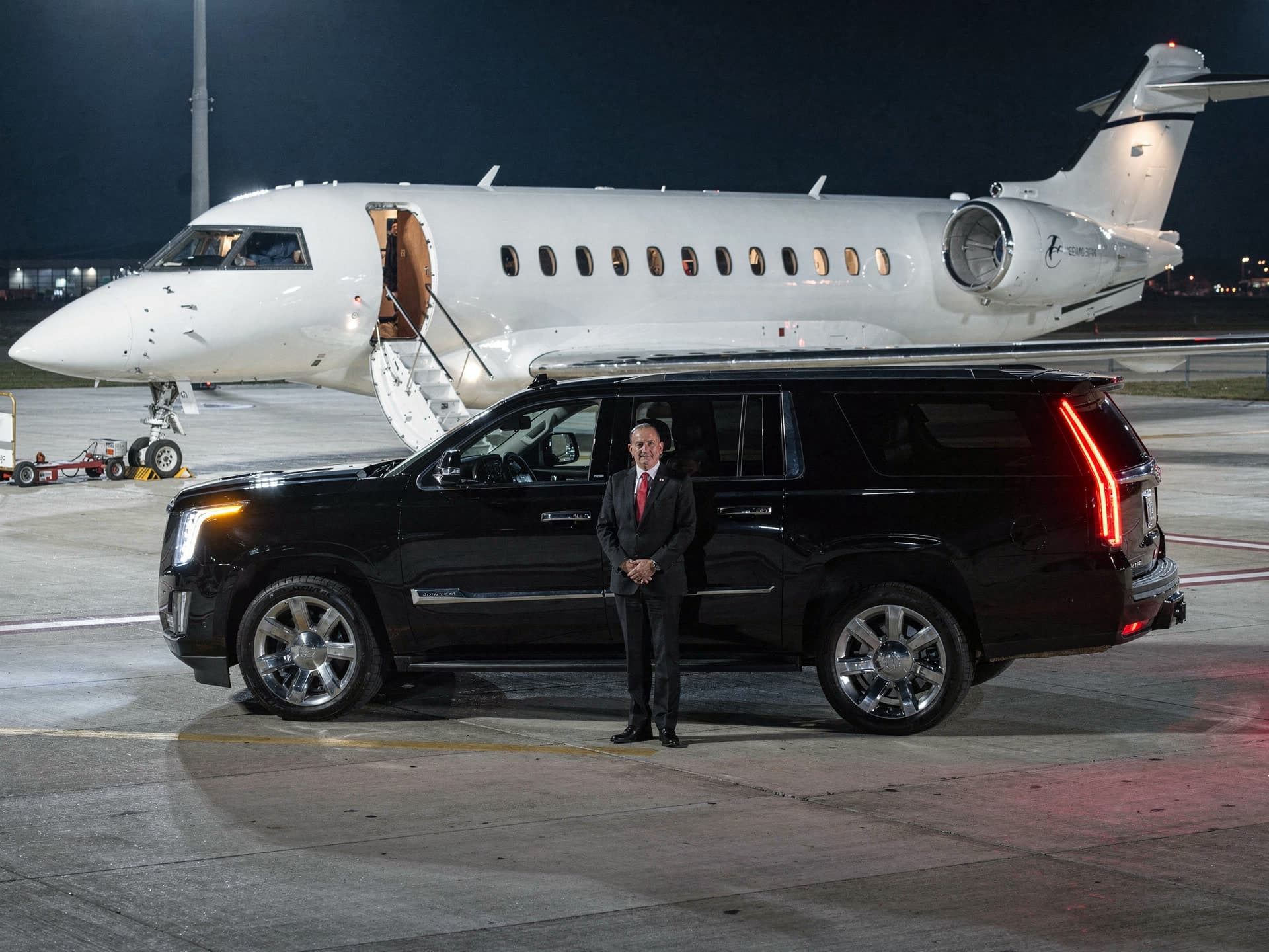 A black Cadillac Escalade parked on a floodlit private terminal ramp at night with its rear door open, a chauffeur in a black suit and red tie standing beside it, and a white business jet with its airstair down behind.