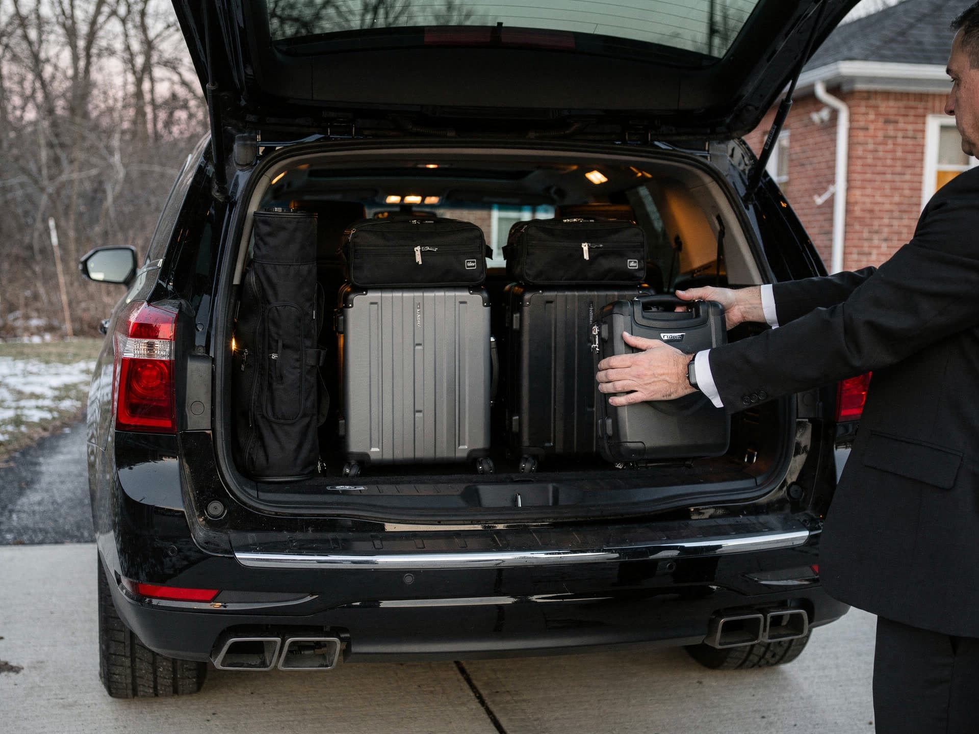 A chauffeur in a black suit sliding a hard shell case into the open rear cargo hold of a black full size SUV in a driveway at winter first light, a long golf bag, two soft carry ons and two more hard cases already packed in behind the rear seats.