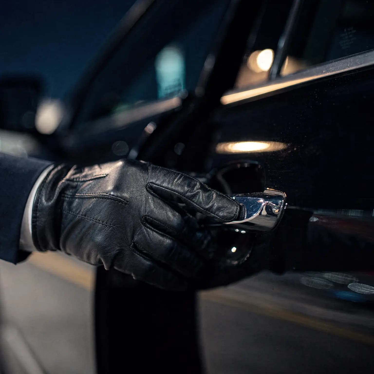 A chauffeur's gloved hand resting on the chrome rear door handle of a black SUV at night, terminal light reflected along the paintwork.