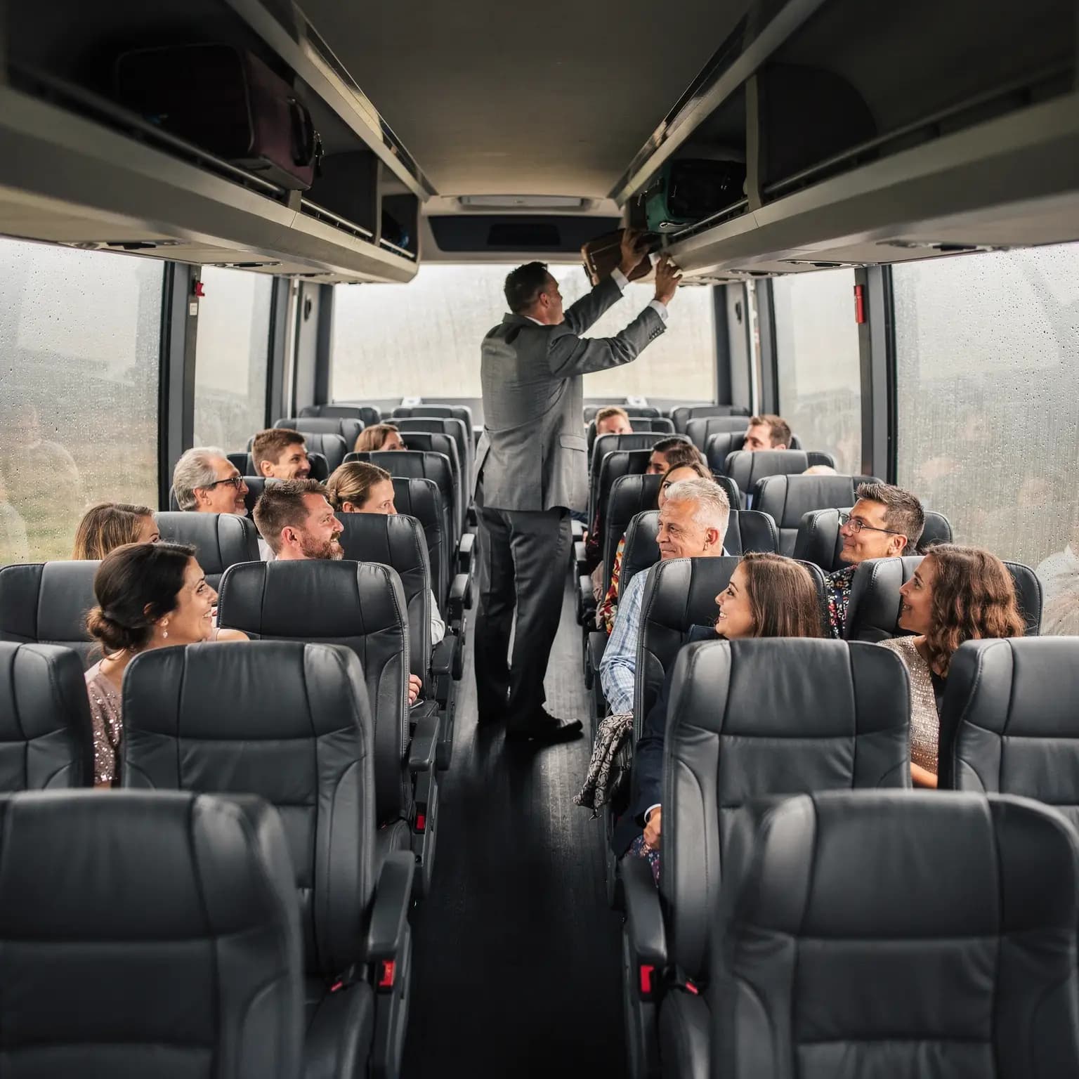 The cabin of a full size motor coach seen from mid aisle, rows of high backed reclining seats running away to the far end with about fourteen passengers in their twenties to seventies turned talking across the aisle, a man in a grey suit standing to slide a weekend bag into the full length overhead rack, rain beading on the panoramic side windows.