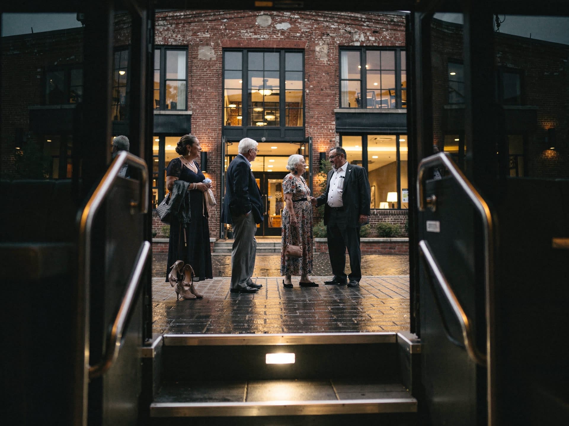 The view out through the open door and lit boarding steps of a motor coach at night, four wedding guests in their sixties and seventies talking on wet brick outside a lit brick mill venue, a pair of blush heels set down on the ground beside one woman in a long dark gown.