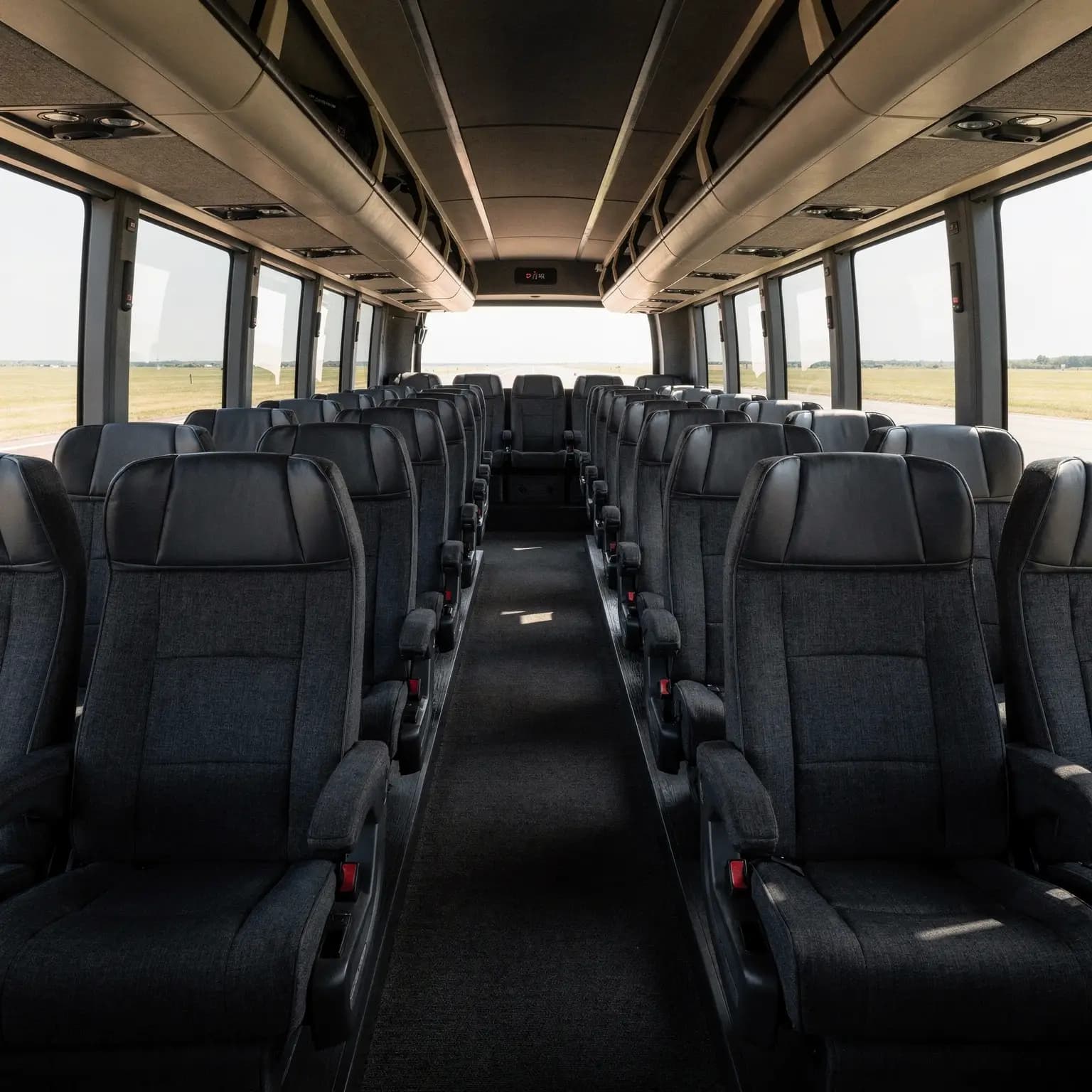 The centre aisle of a full size motor coach, rows of high backed reclining seats and full length overhead parcel racks running away toward the front of the cabin.