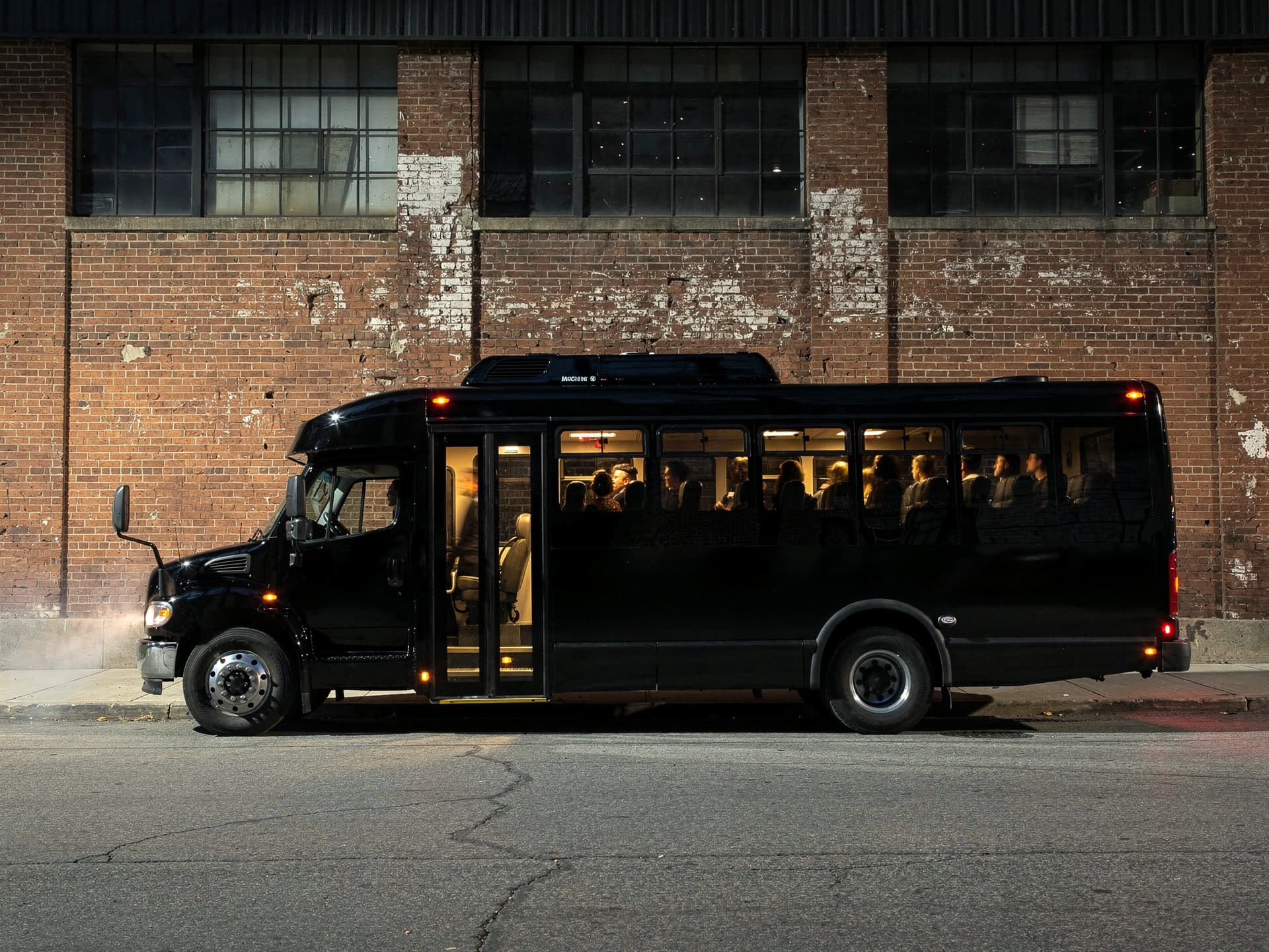 A black party bus photographed side on against a weathered brick mill wall at night, its boarding door open on a lit stairwell and its warm lit cabin full of seated passengers visible through the side windows.