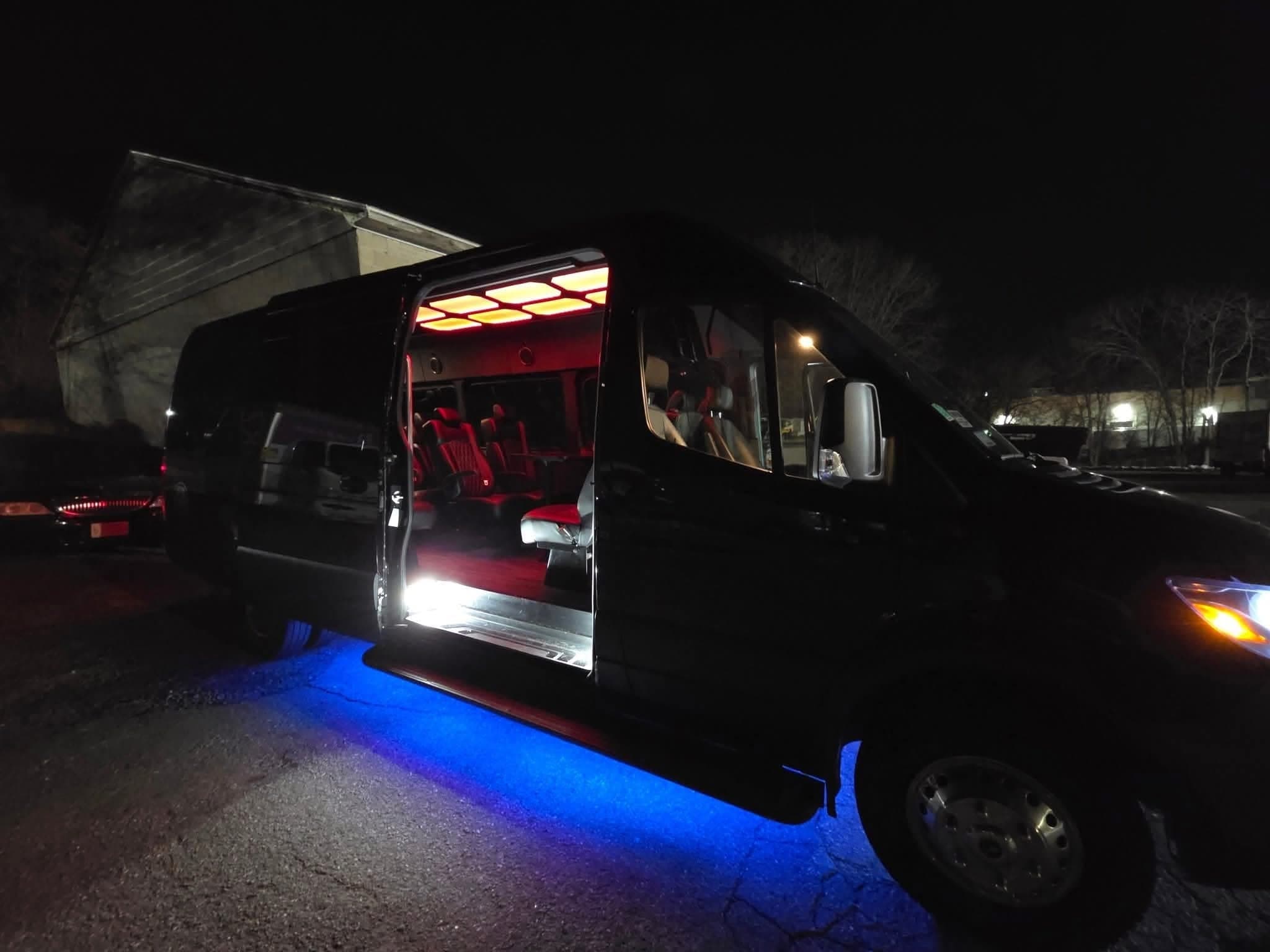 A black Mercedes Sprinter parked at night with its side door slid fully open, revealing a walk in cabin lit in red with quilted red leather seating, a honeycomb ceiling light panel and an illuminated boarding step, blue accent light spilling onto the pavement below the sill.