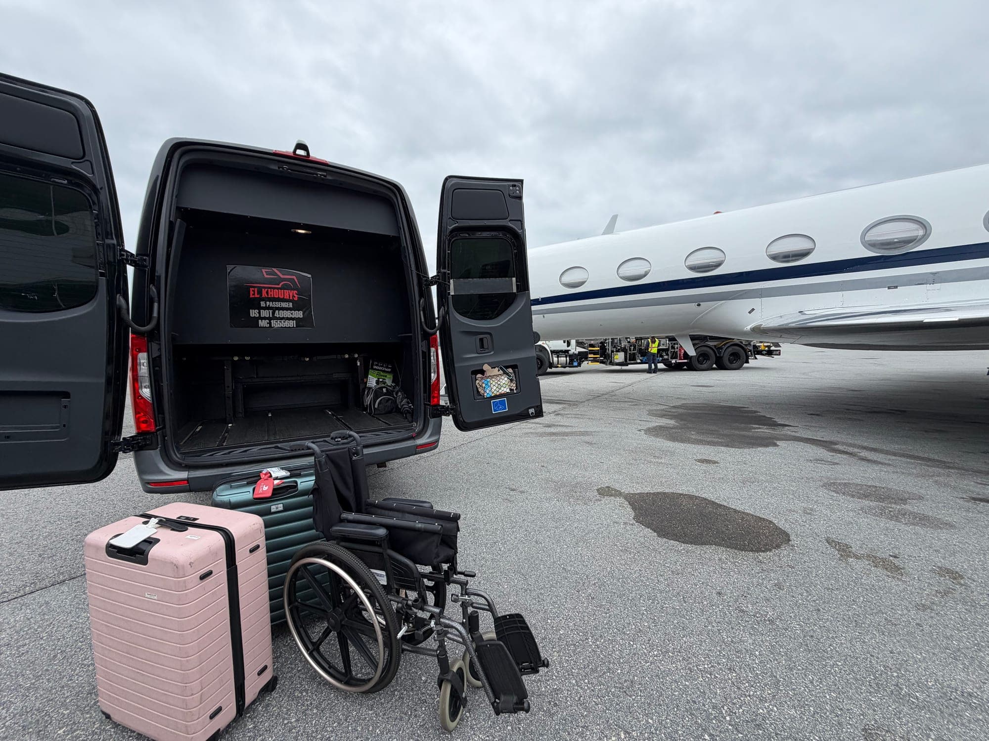 The rear of a black El Khourys Sprinter on an airport tarmac with both rear doors open beside a parked private jet, a pink hardshell suitcase, a teal roller bag and a folded wheelchair waiting on the ground, a US DOT and MC compliance decal on the inside of the door.