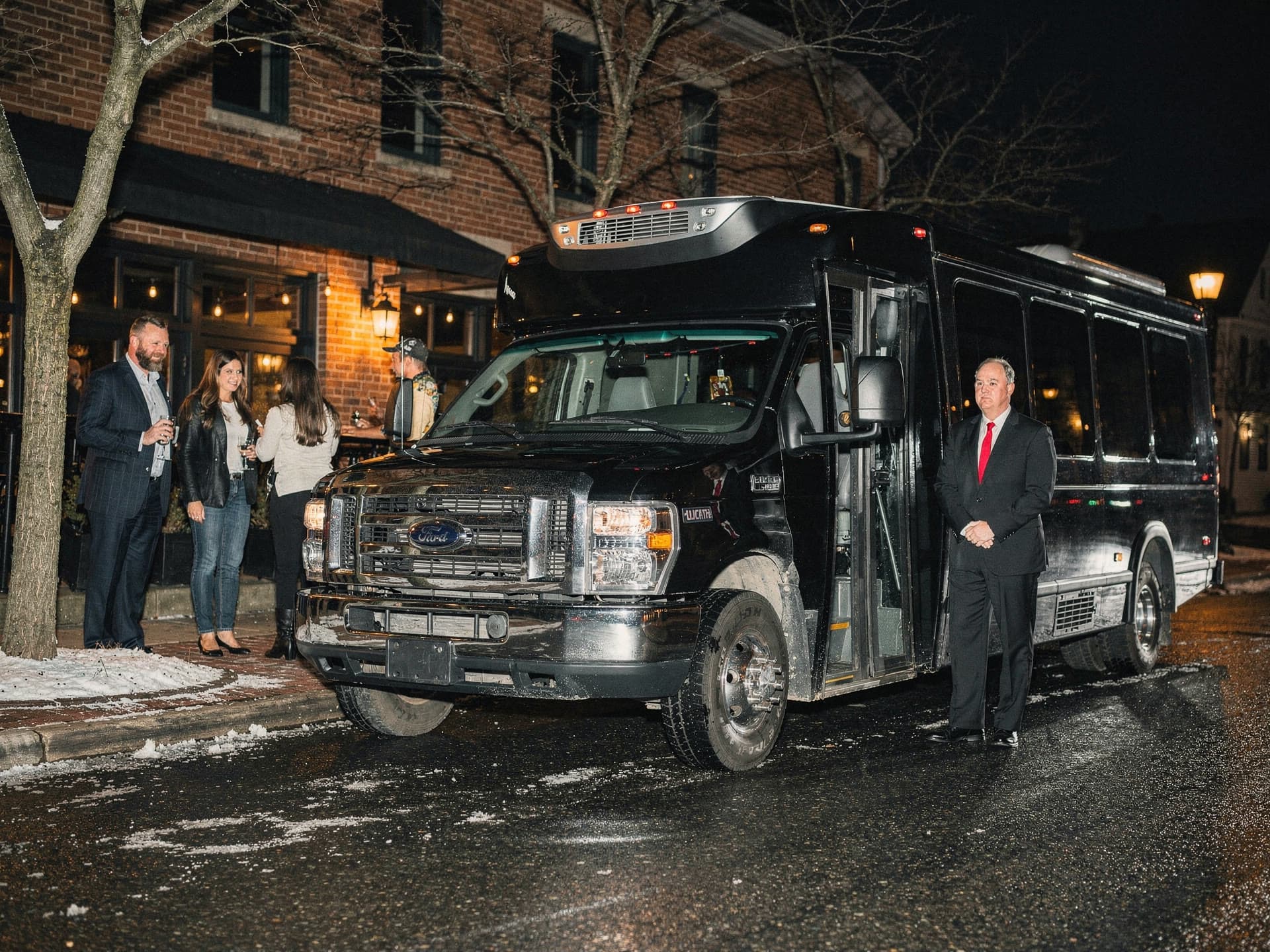 A compact black party bus parked outside a restaurant at night, interior lighting glowing through tinted windows, a small group boarding.