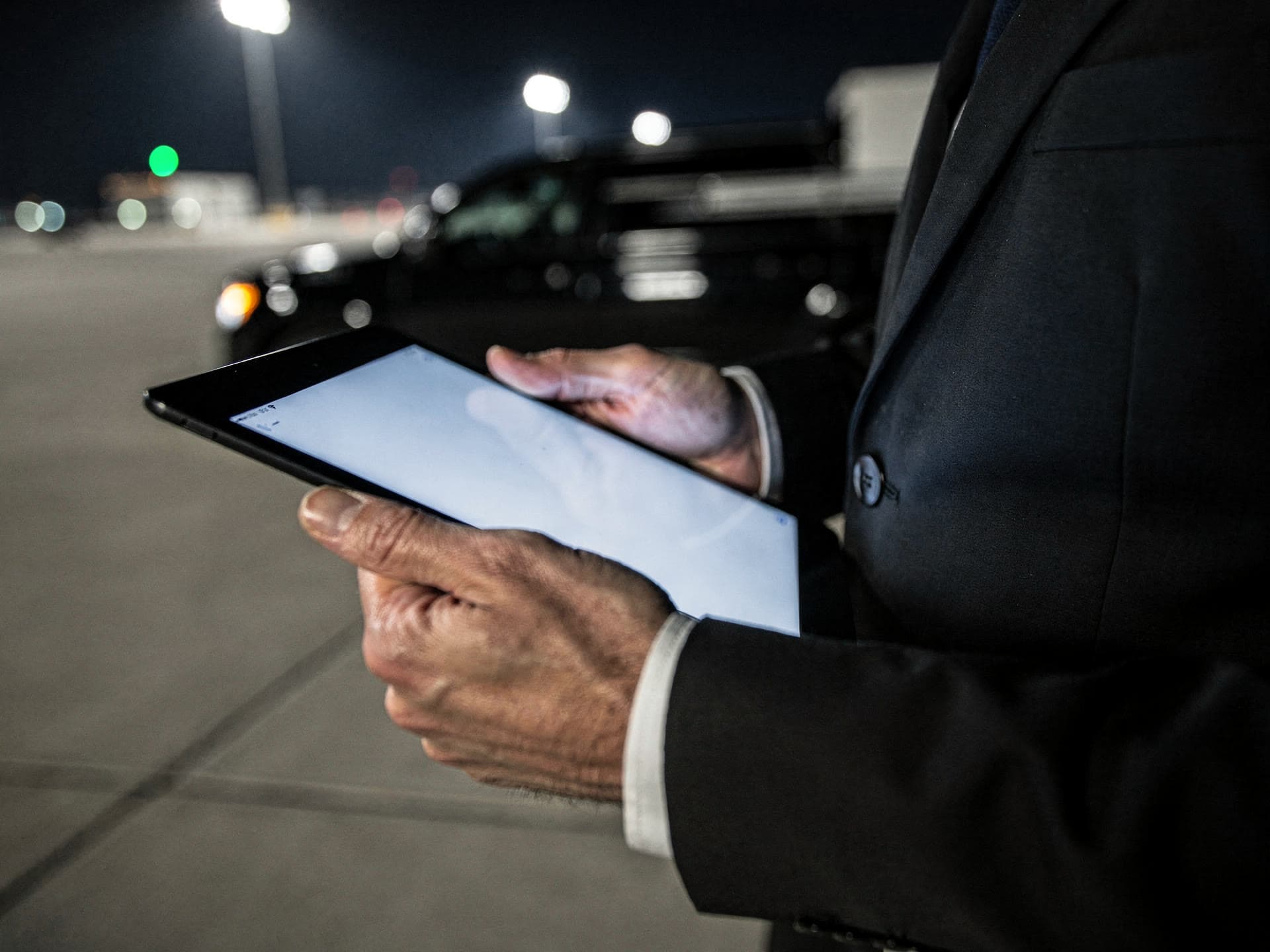 A chauffeur's hands holding a tablet low at night, the screen glowing, ramp lights out of focus behind it.