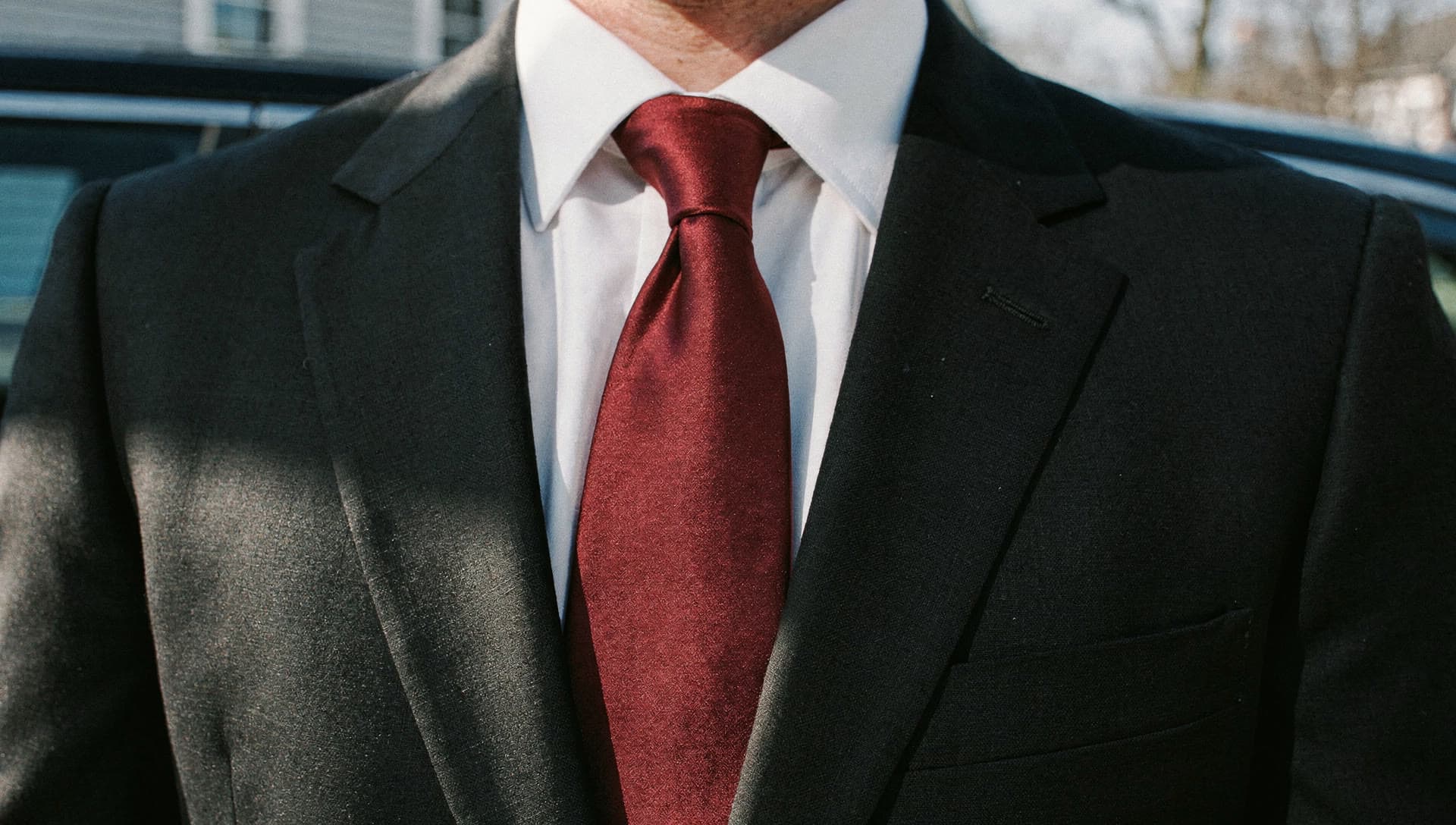 A red tie against a black suit jacket in daylight, a vehicle out of focus behind