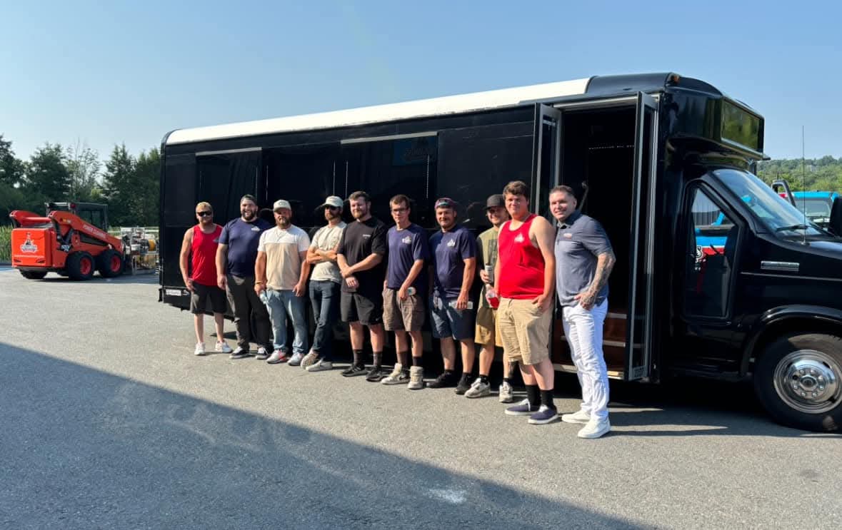 Eleven men in work clothes lined up shoulder to shoulder beside a black bus with its door open, on a paved lot in summer daylight, a compact loader parked behind them.