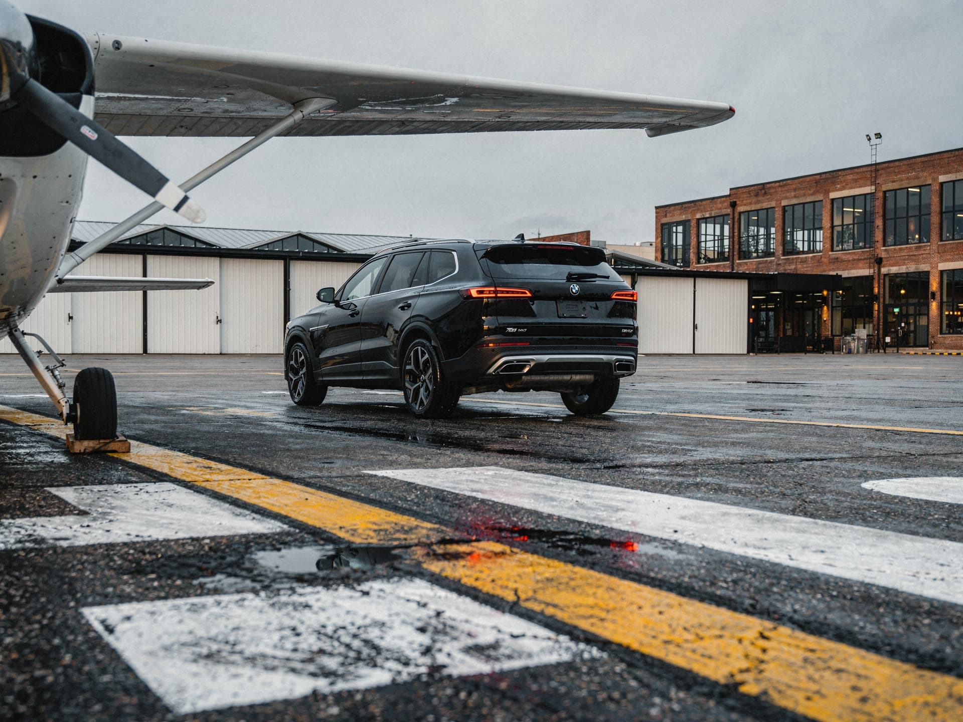 Seen from inside a hangar past the leading edge of a light aircraft wing, a black SUV waits outside in daylight.