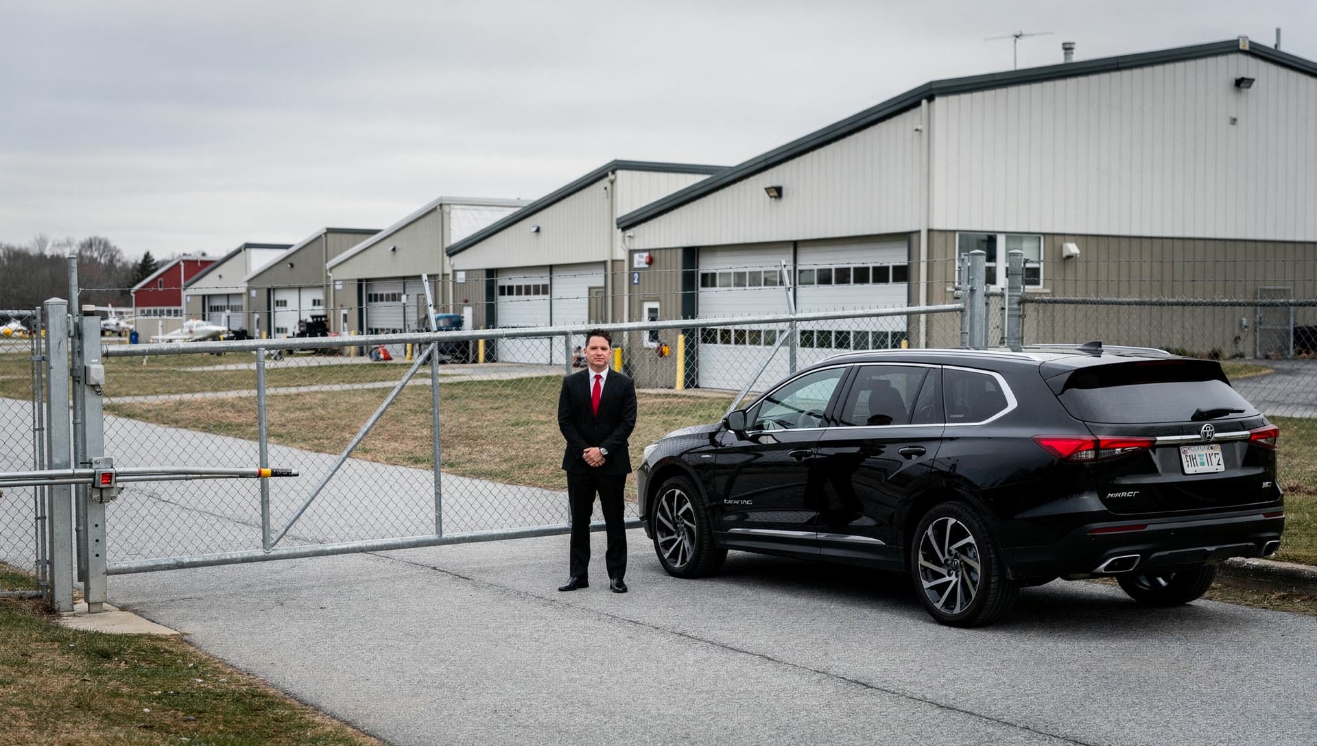 A general aviation perimeter gate and hangar row in daylight, a black SUV waiting on the access road with a chauffeur standing beside it.