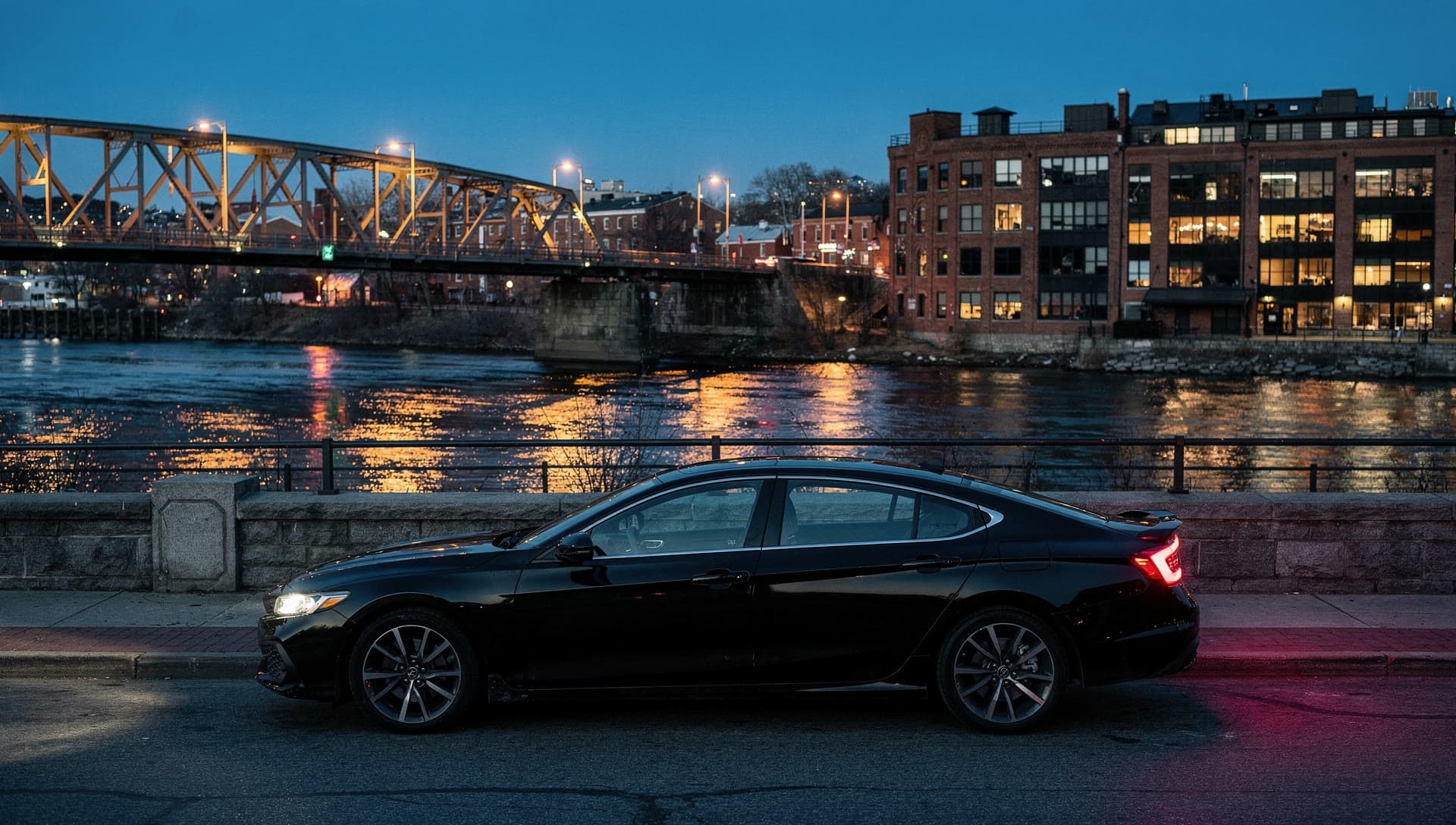 A car at a Haverhill riverfront curb at blue hour with the lit Merrimack bridge span behind it.