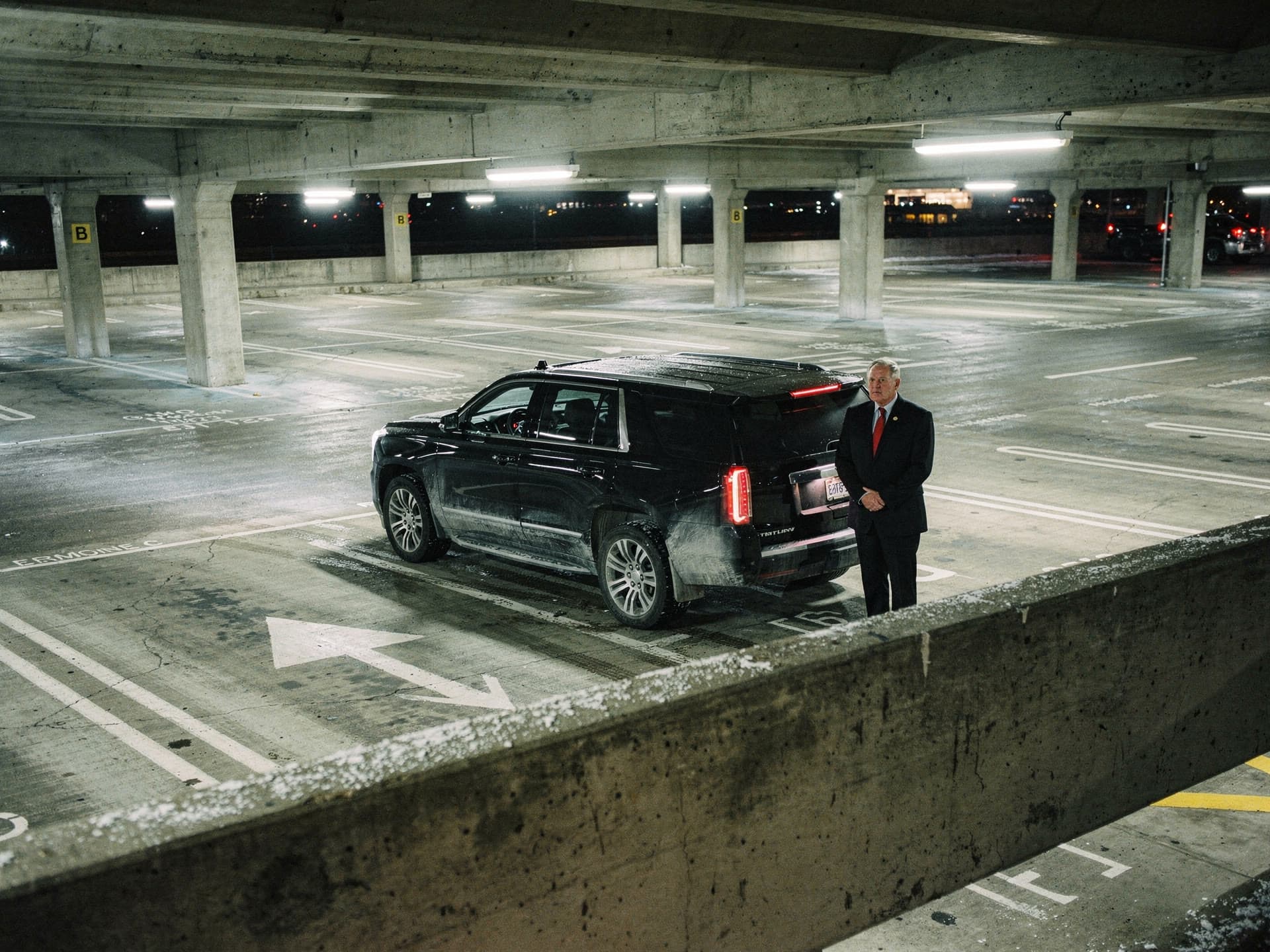 A high overview at night looking down from an upper level of an airport parking garage at the marked limousine stand lane below, one black SUV waiting in it, terminal light beyond.