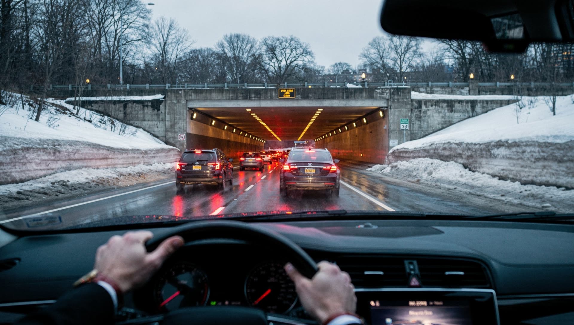 The approach to a harbour tunnel portal at first light seen through a windshield, wet road, tail lights ahead, the driver's hands at the bottom of the frame.