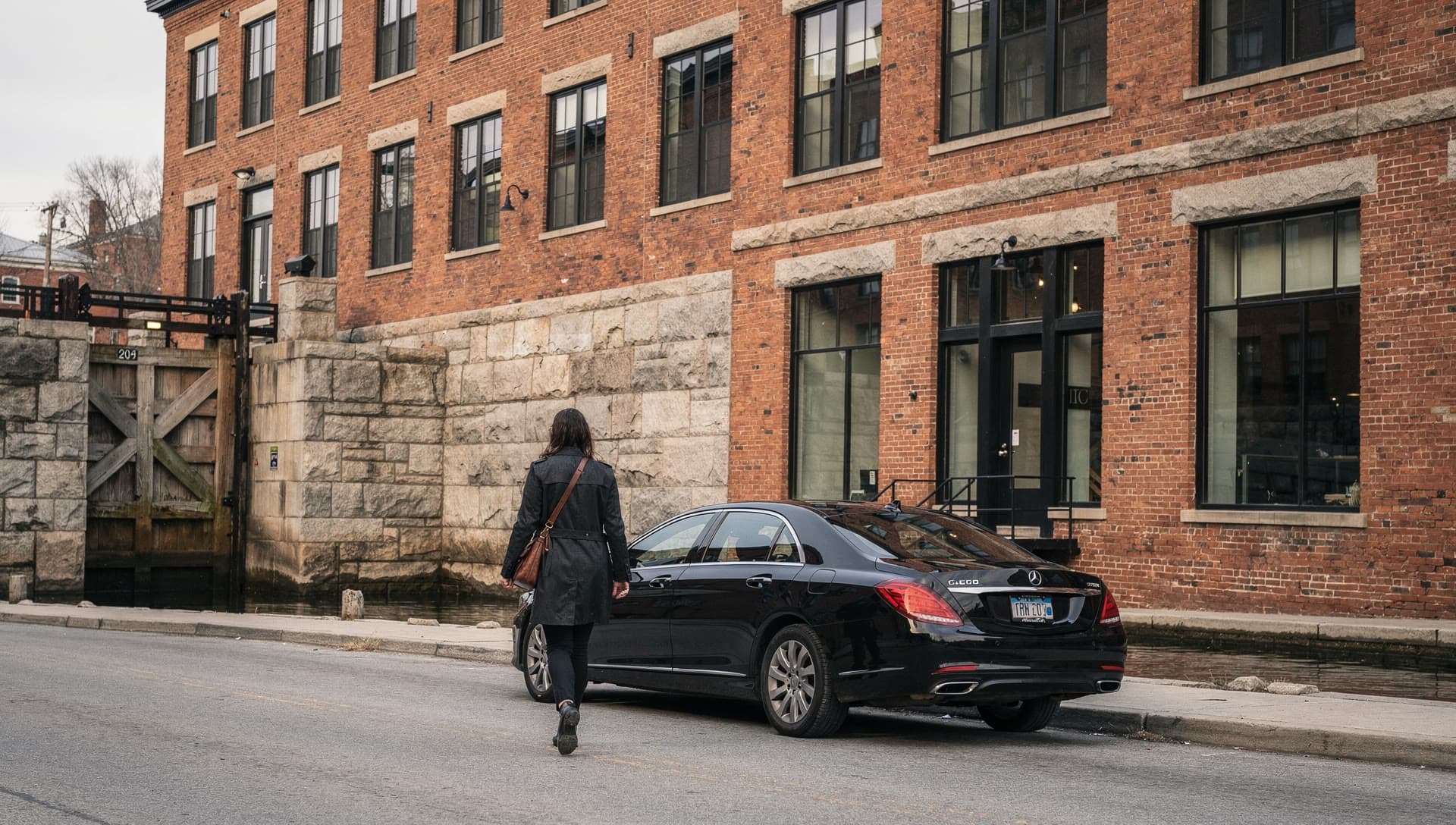 A passenger walking toward a waiting car past a canal lock and mill brick in Lowell on a mid morning.