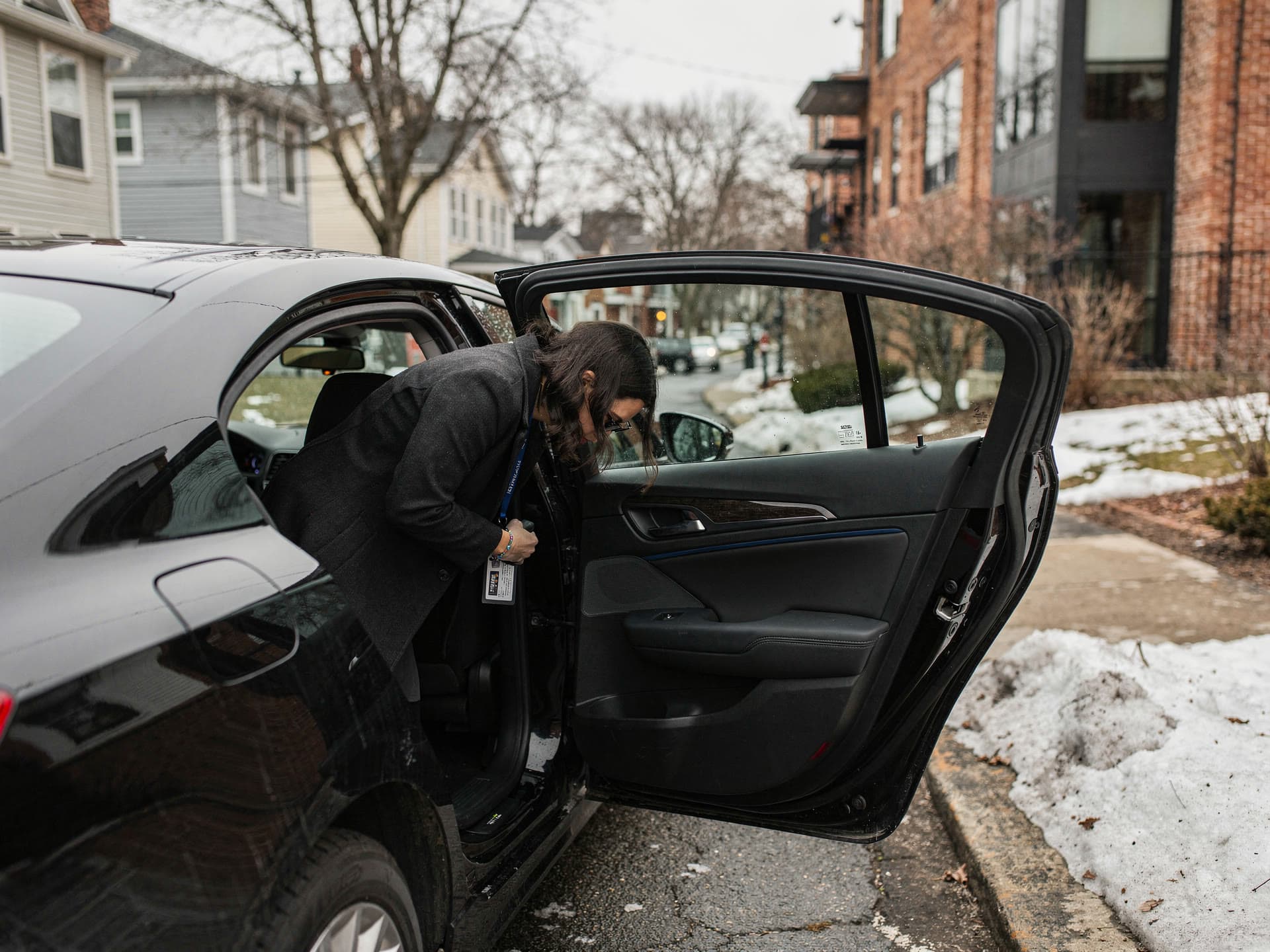 A professional in a coat ducking into the back seat at the curb, seen over the shoulder from behind the open rear door.