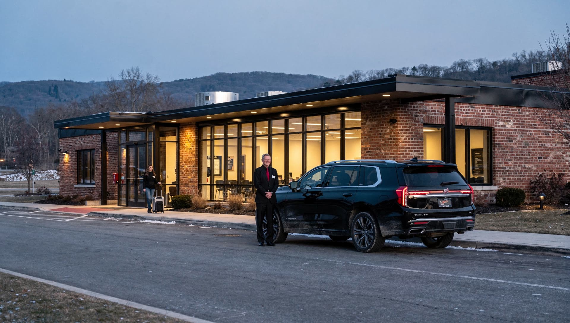 A small regional airport terminal frontage at dusk with an SUV waiting at a nearly empty kerb and low hills on the horizon behind it.