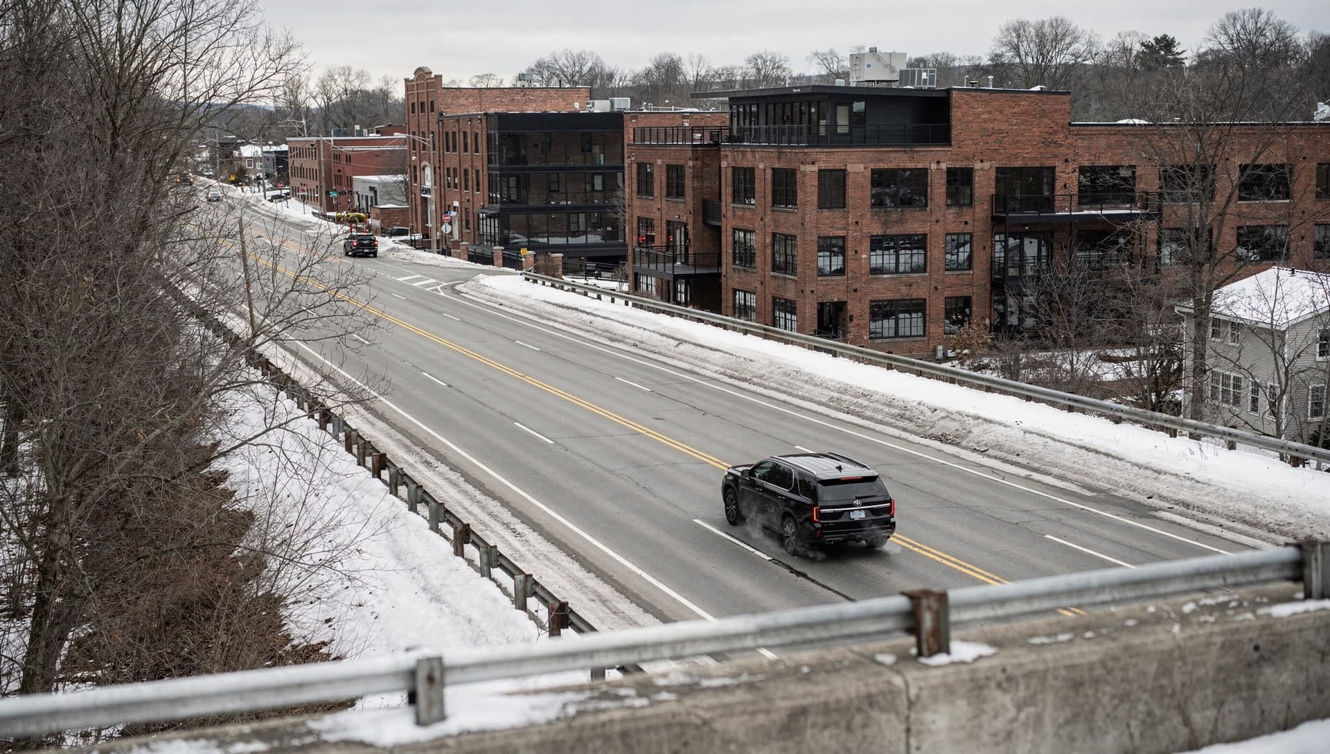 Seen from a highway overpass, a single black SUV runs the near lane of a divided road past restored brick mill buildings, with plowed snow banked along the guardrails and bare winter trees on both sides.