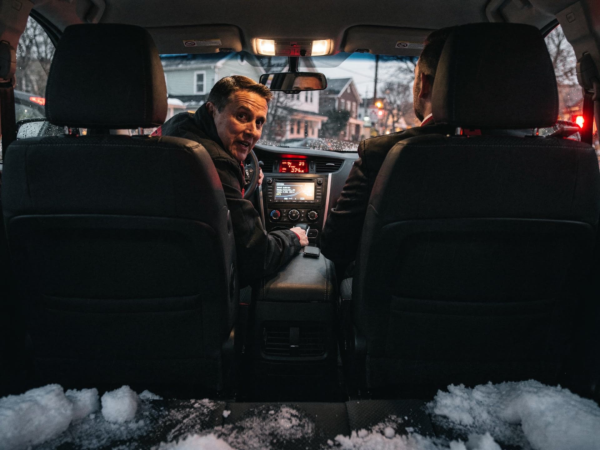 A rider leaning forward between the front seats to speak to the chauffeur while the car waits at a light, city glow through the glass.