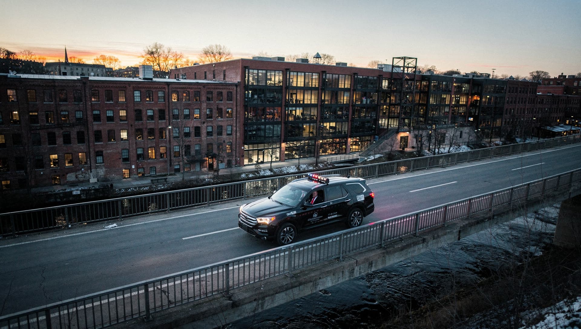 An SUV crossing a bridge above the Amoskeag millyard in Manchester, New Hampshire at dusk, the river below.