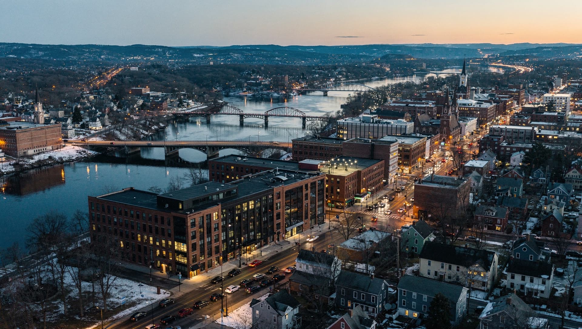 The Merrimack Valley at dusk seen from above, mill town rooftops and river bridges with the lights coming on.