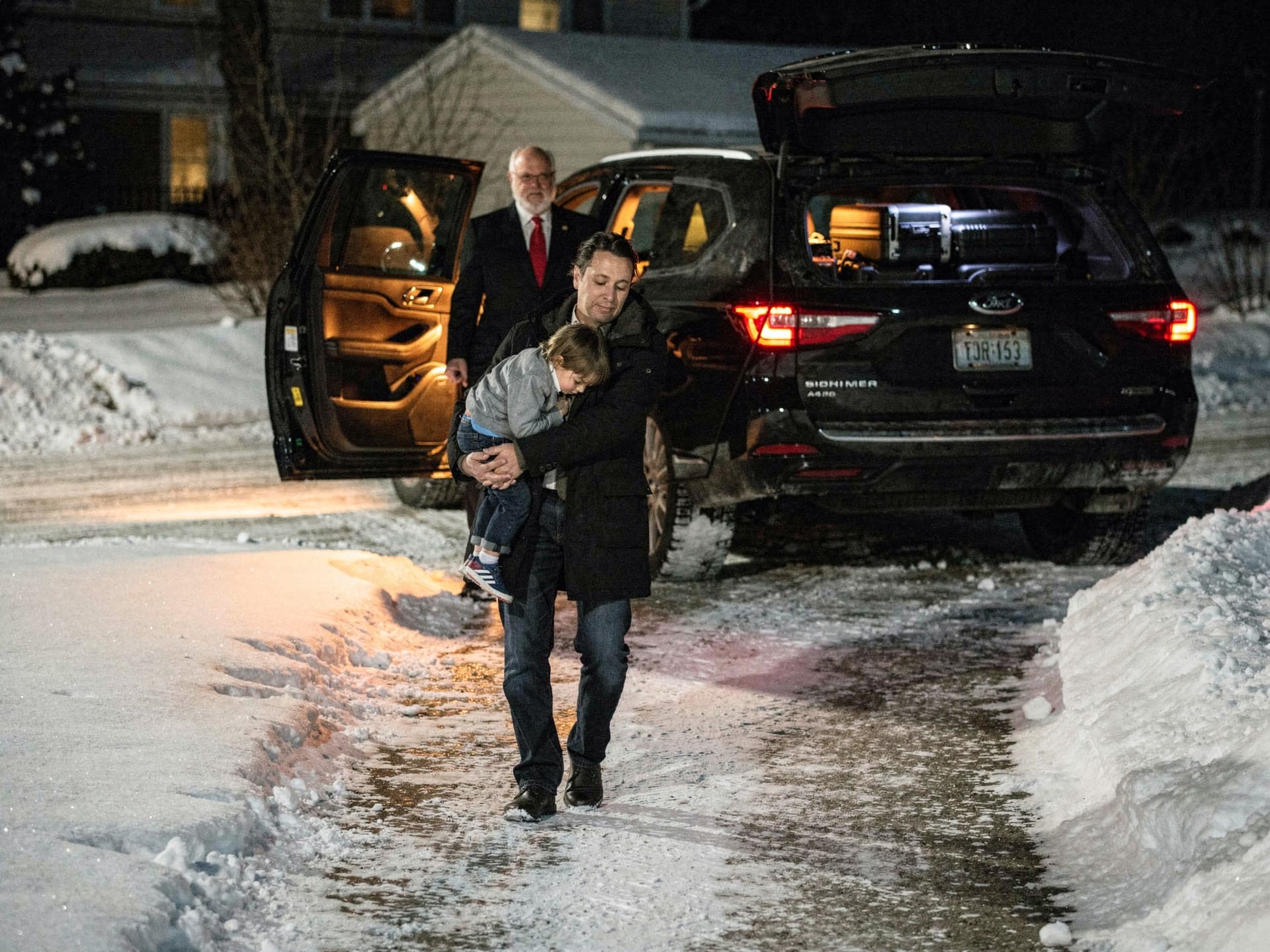 A parent carrying a sleeping child to the open rear door of an SUV in the dark, cases already loaded behind them.