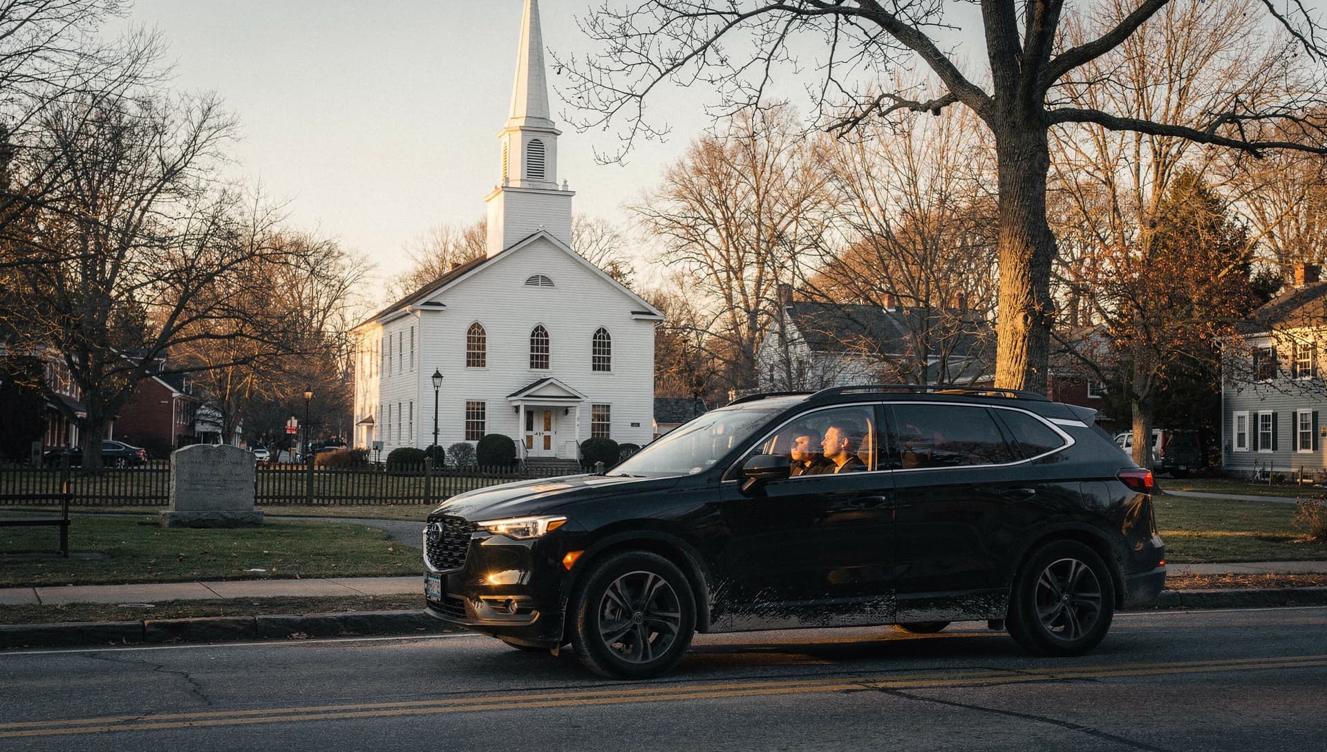 An SUV on the road past the North Andover town common at golden hour, a white clapboard church and steeple behind it.