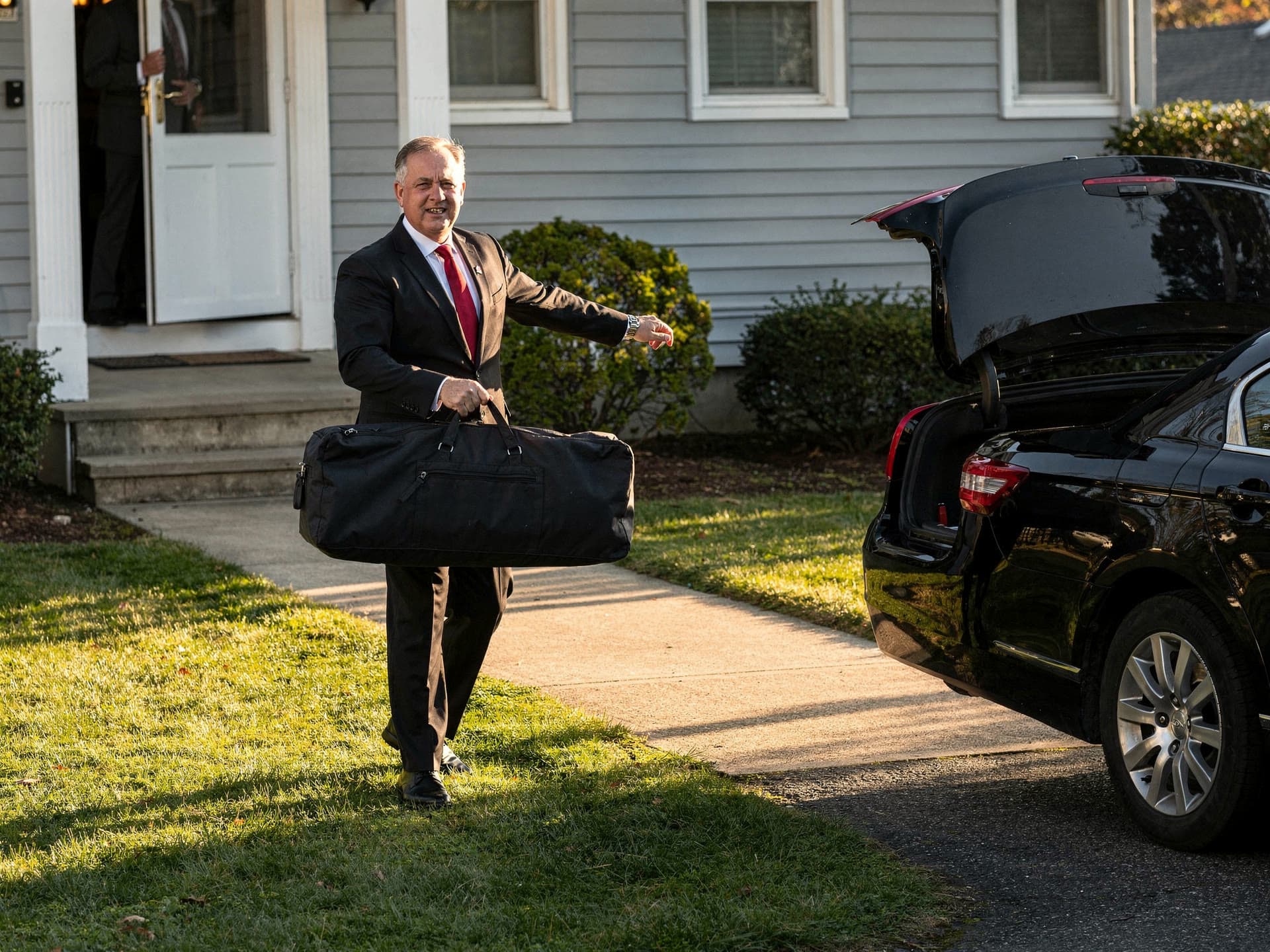 A chauffeur carrying a garment bag flat over one arm from a front door to an open trunk in the late afternoon.