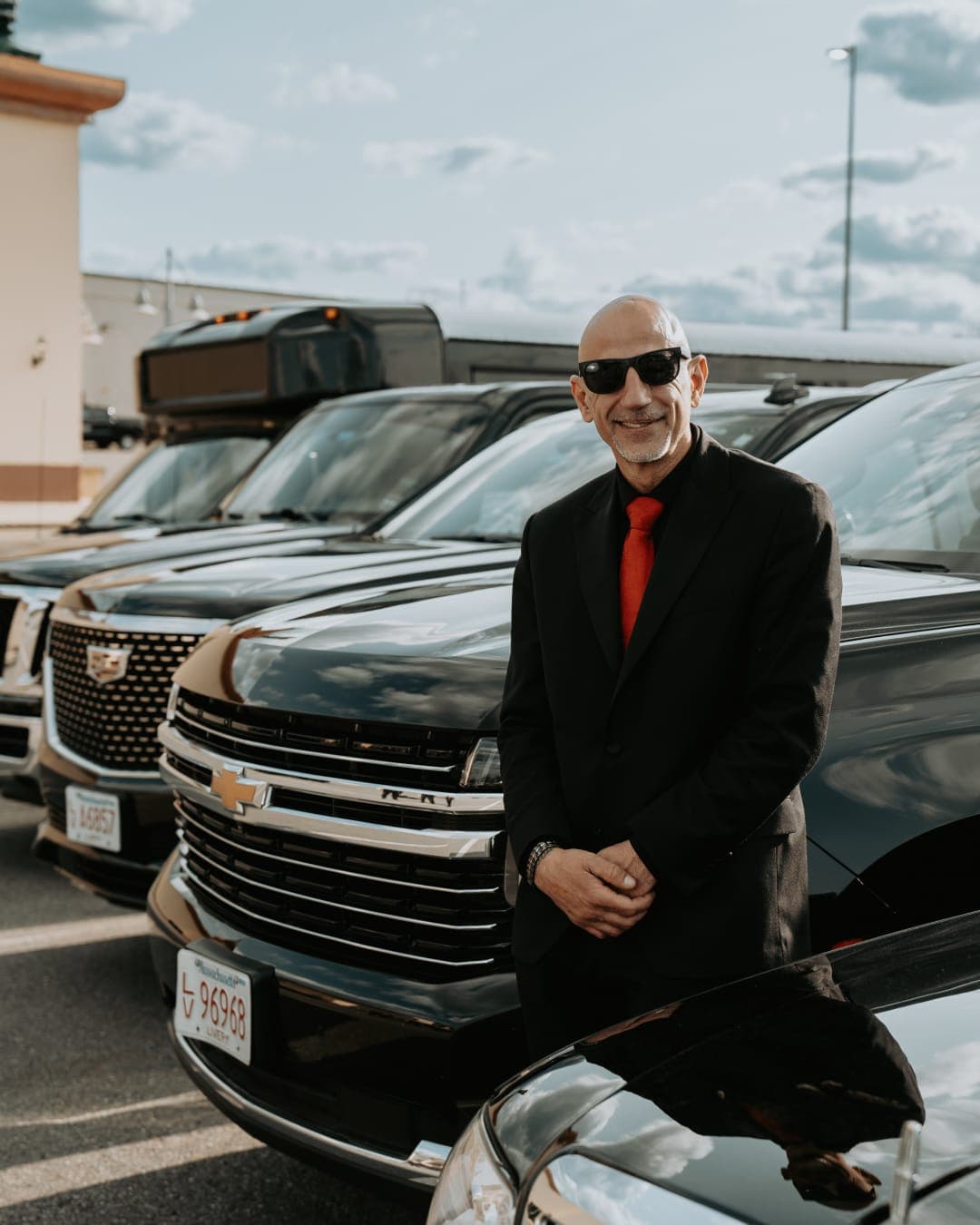 Hanna Elkhoury in a black suit and red tie standing in front of his own Escalade and Tahoe