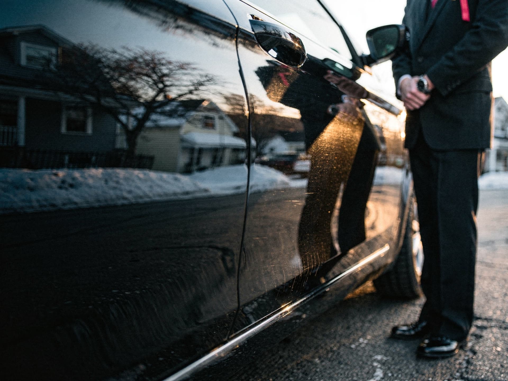 A close view of a chauffeur straightening a red tie, reflected in the polished black door panel of a vehicle in early morning light.